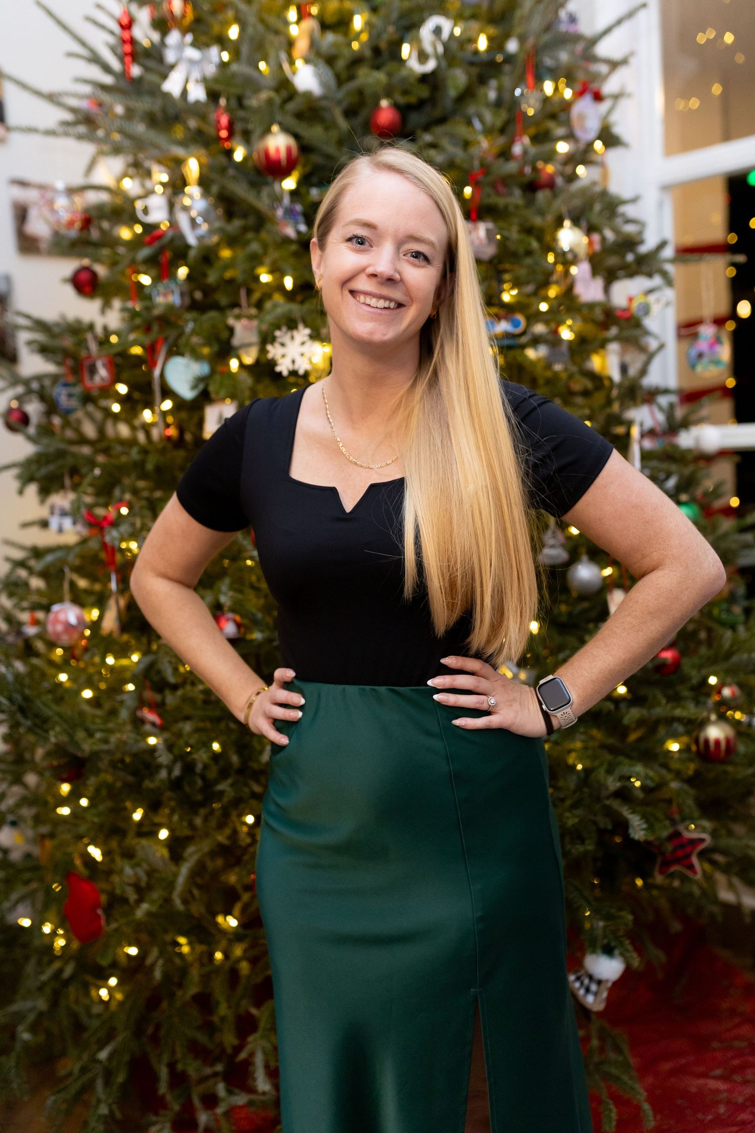 Woman in black top and dark green skirt poses in front of decorated Christmas tree.