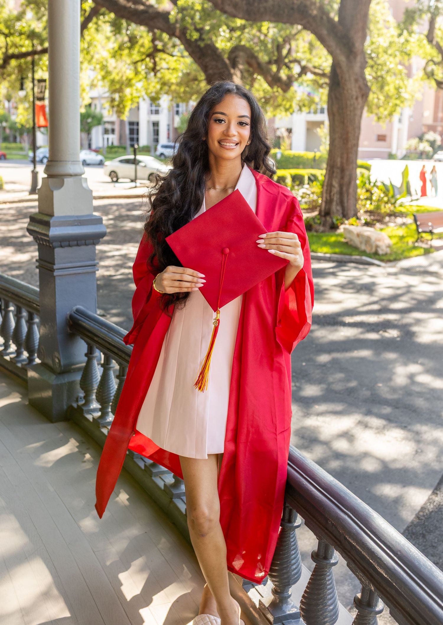 Young woman in red graduation gown and cap smiling on campus steps, celebrating her graduation day.