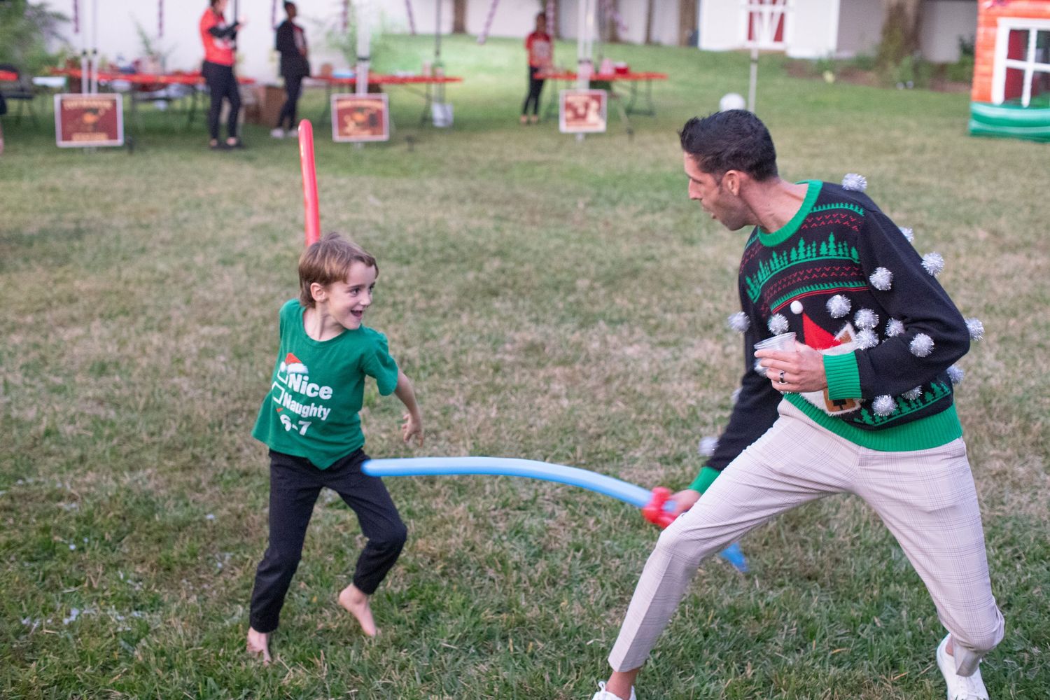 Two people play an outdoor game with a pool noodle while wearing festive holiday sweaters.