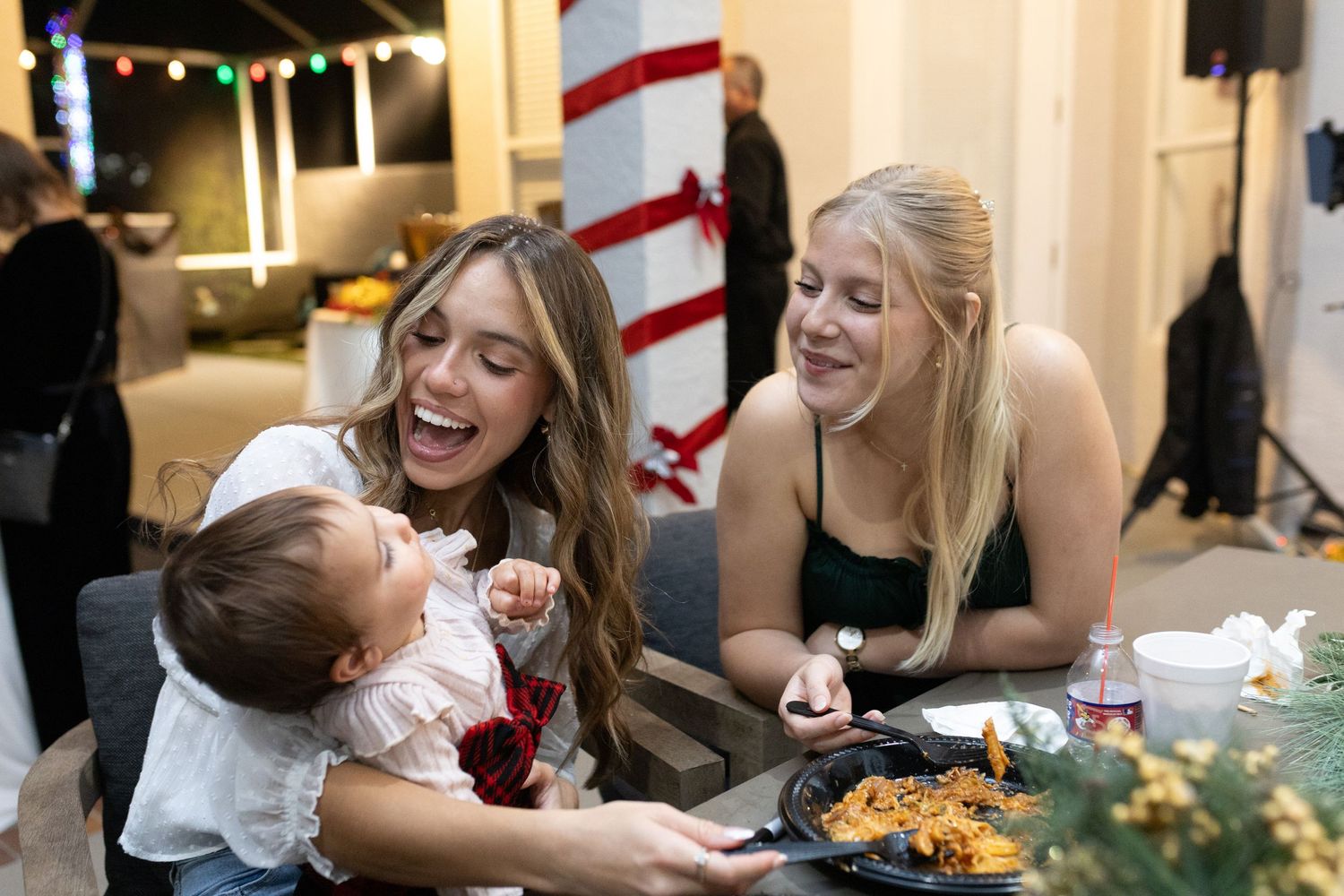 People sharing laughter and joy during a holiday meal with decorative lights in the background.