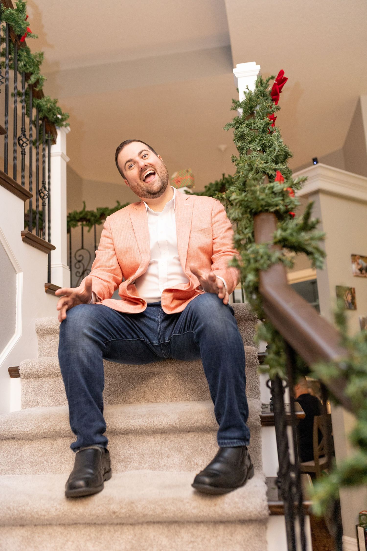 Someone wearing a peach-colored blazer and jeans sits on carpeted stairs decorated with holiday greenery.