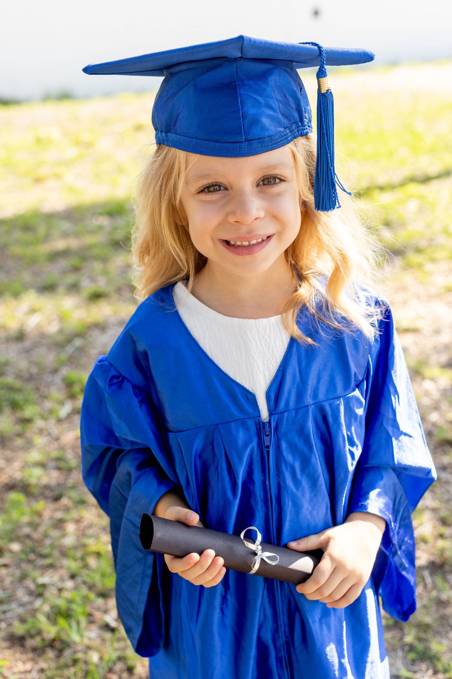 Young graduate in bright blue cap and gown holding diploma while smiling outdoors.