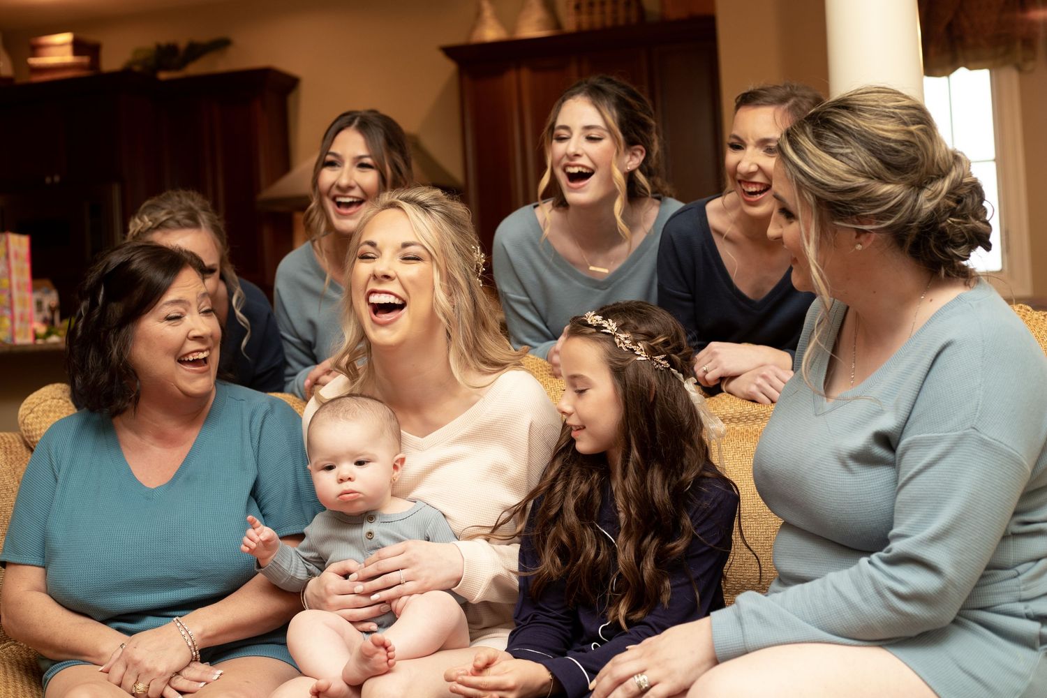 Bride laughing with bridesmaids, family, and a baby girl wearing a floral crown during a joyful wedding preparation gathering.