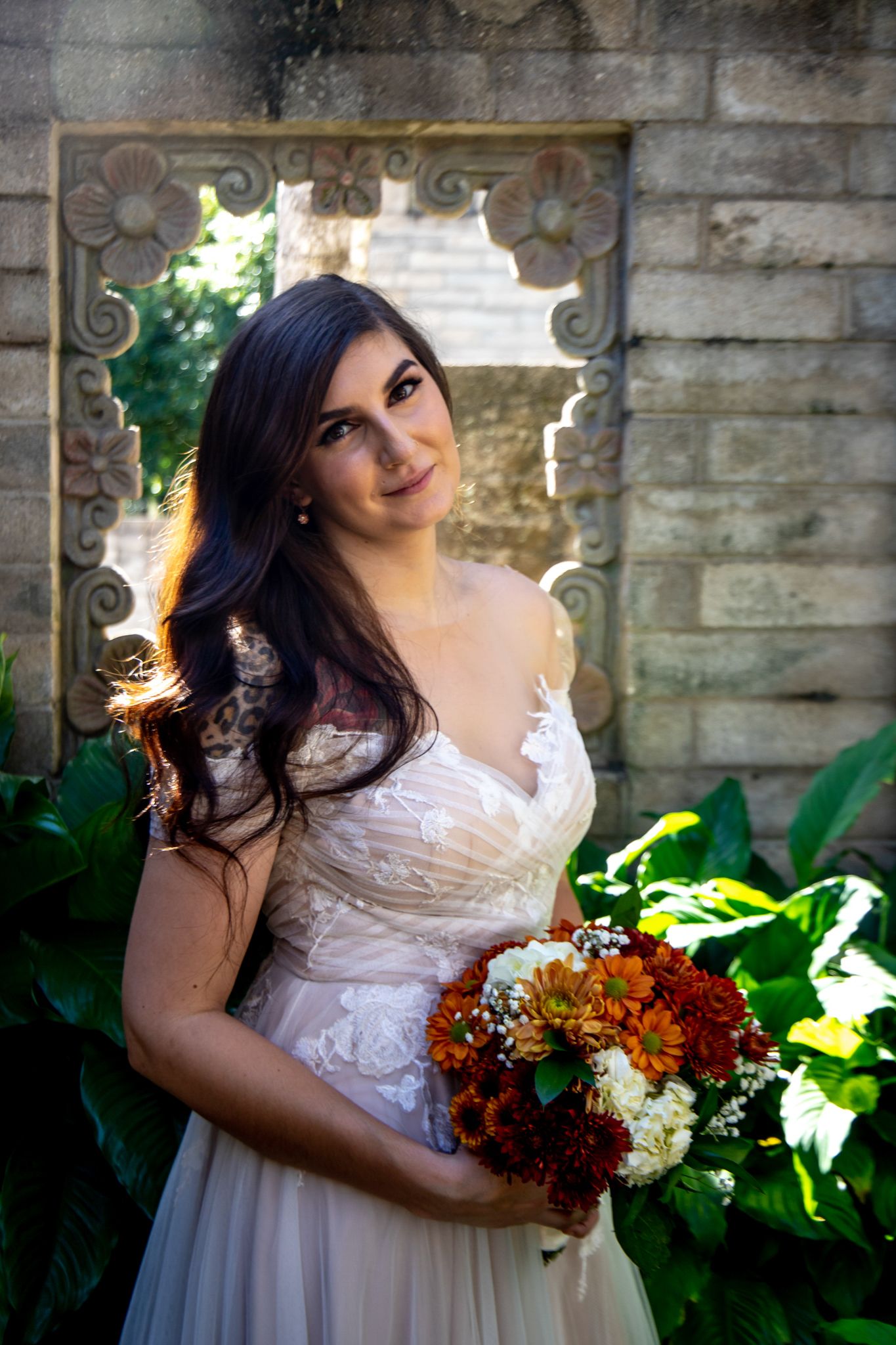 Bride in lace dress poses with autumn-colored bouquet against stone wall backdrop.