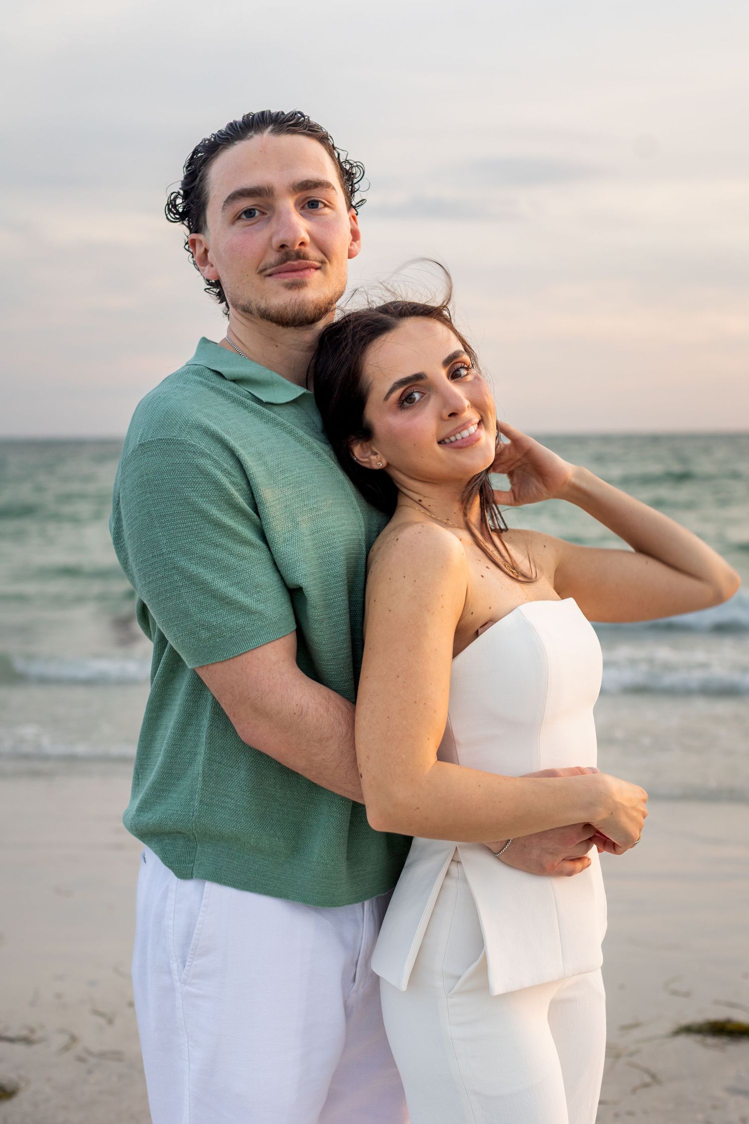 A couple embraces on a beach at sunset, man in green shirt and woman in white outfit, smiling at camera.
