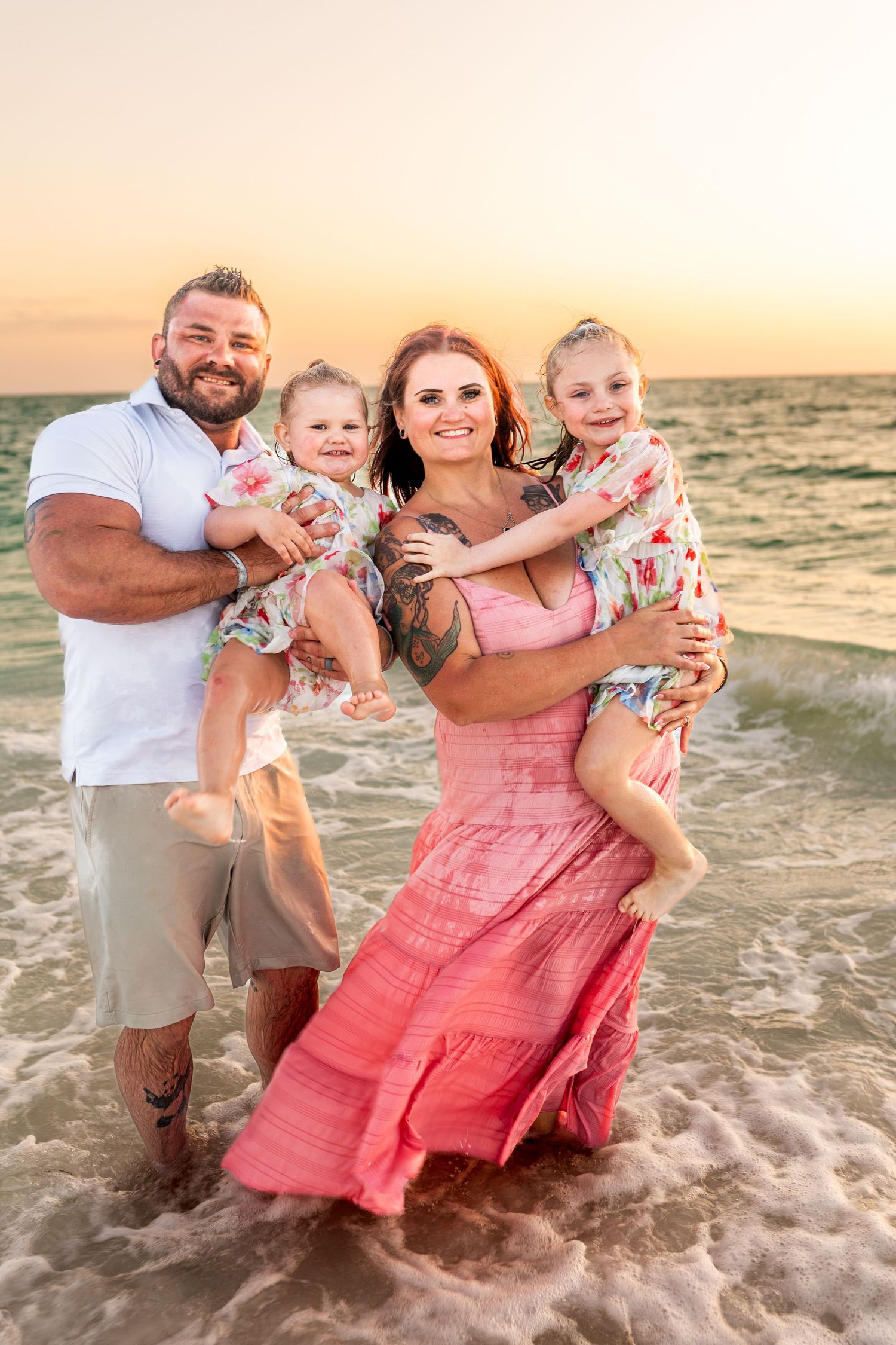 Family of four posing at sunset beach, parents holding two young girls in matching floral outfits, waves at their feet.