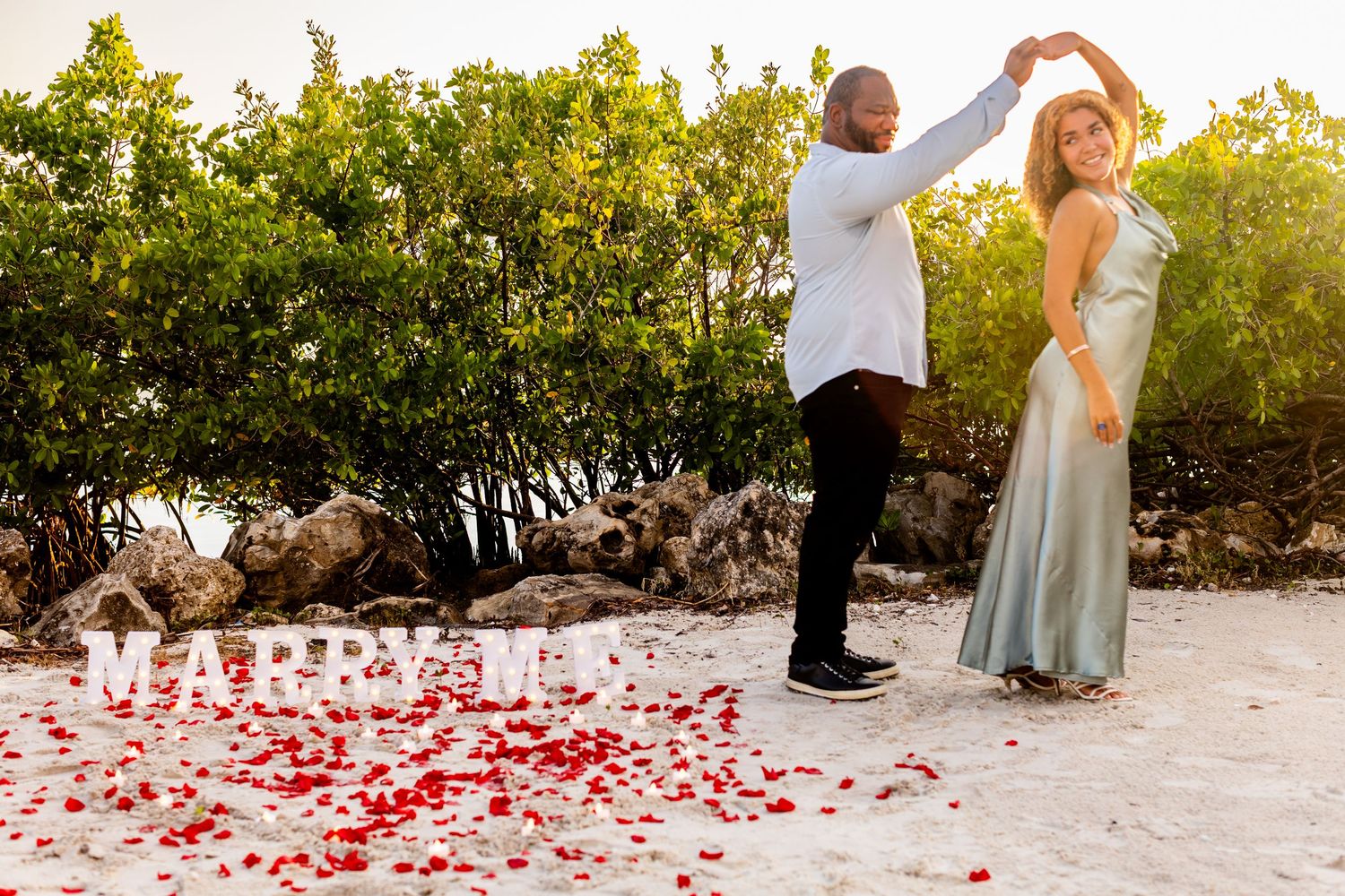 Couple dancing on beach with rose petals and Marry Me sign during romantic outdoor proposal at sunset.