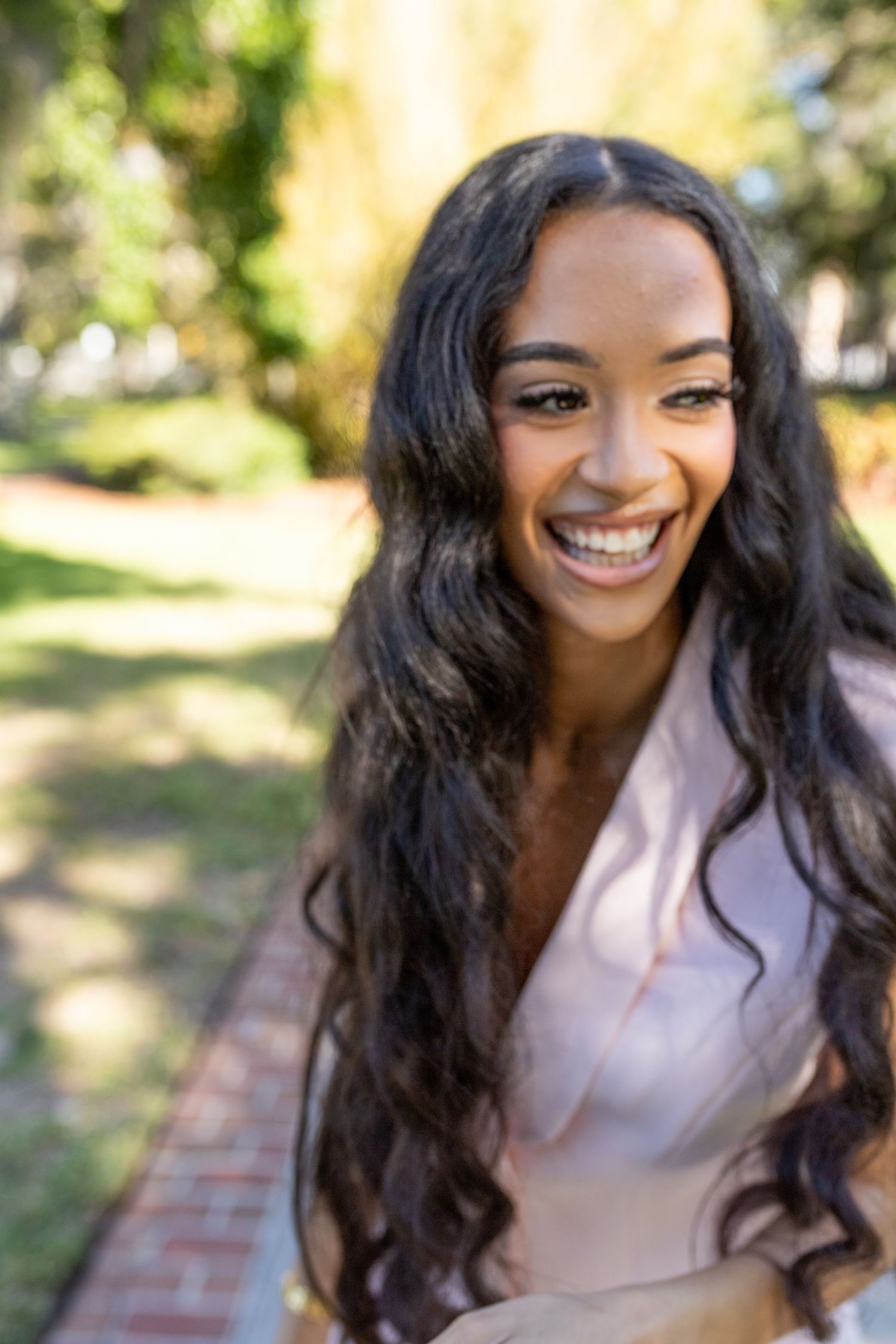 Smiling young woman with long wavy dark hair standing outdoors in a sunny park, wearing a light pink top.