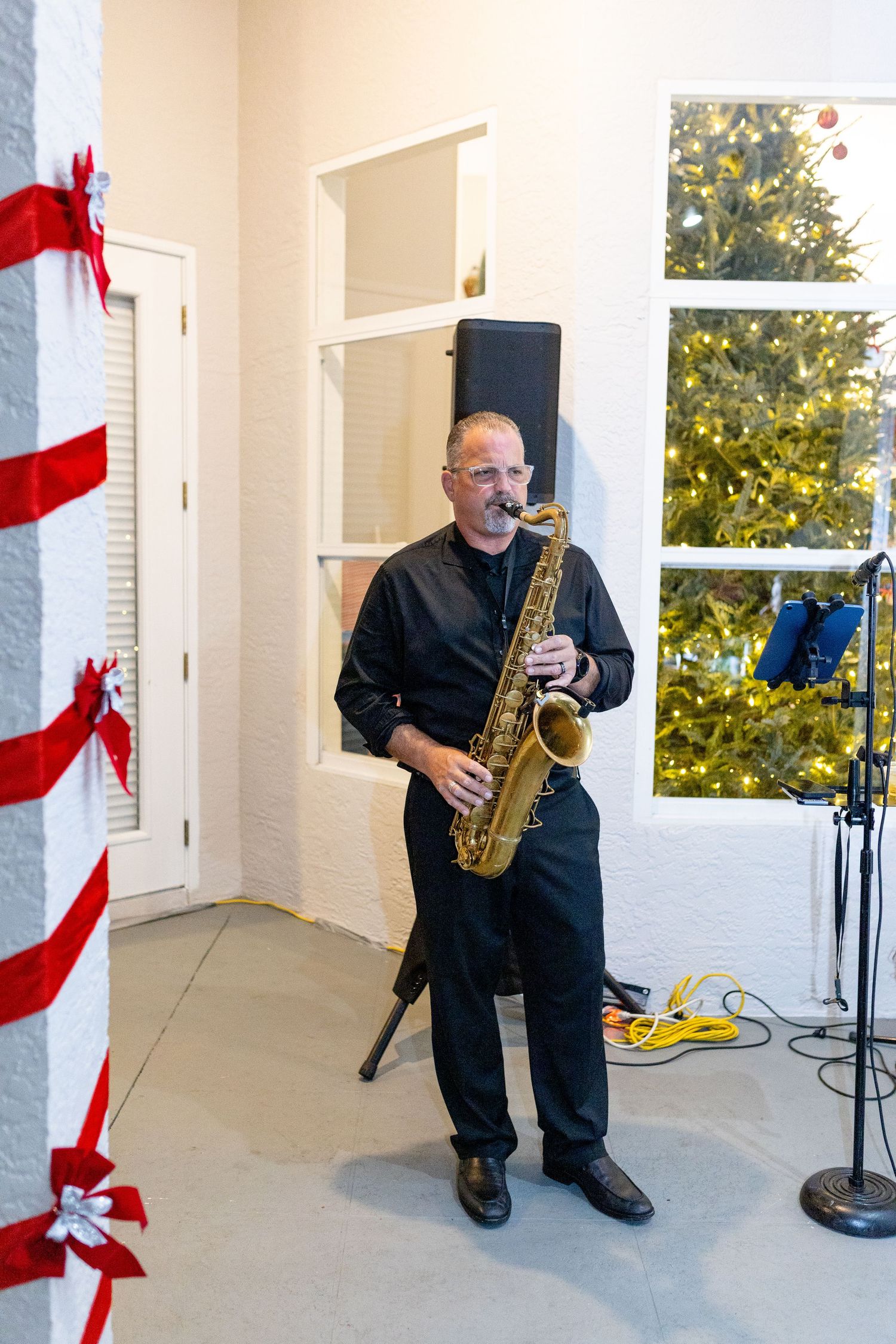 A musician plays saxophone during a holiday performance on a decorated porch with red and white accents.