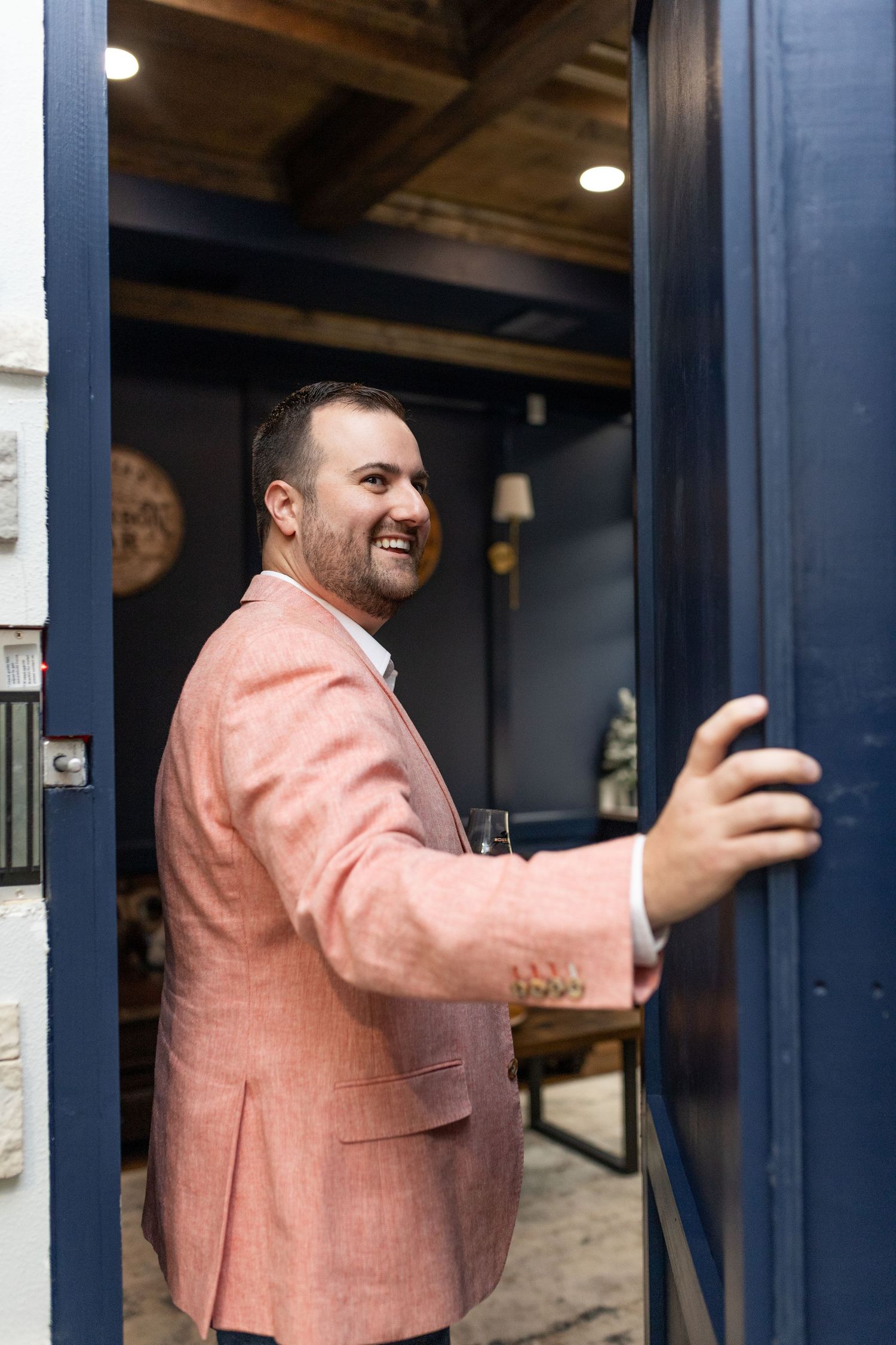 A person in a salmon-colored blazer smiles while opening a blue door at an establishment.