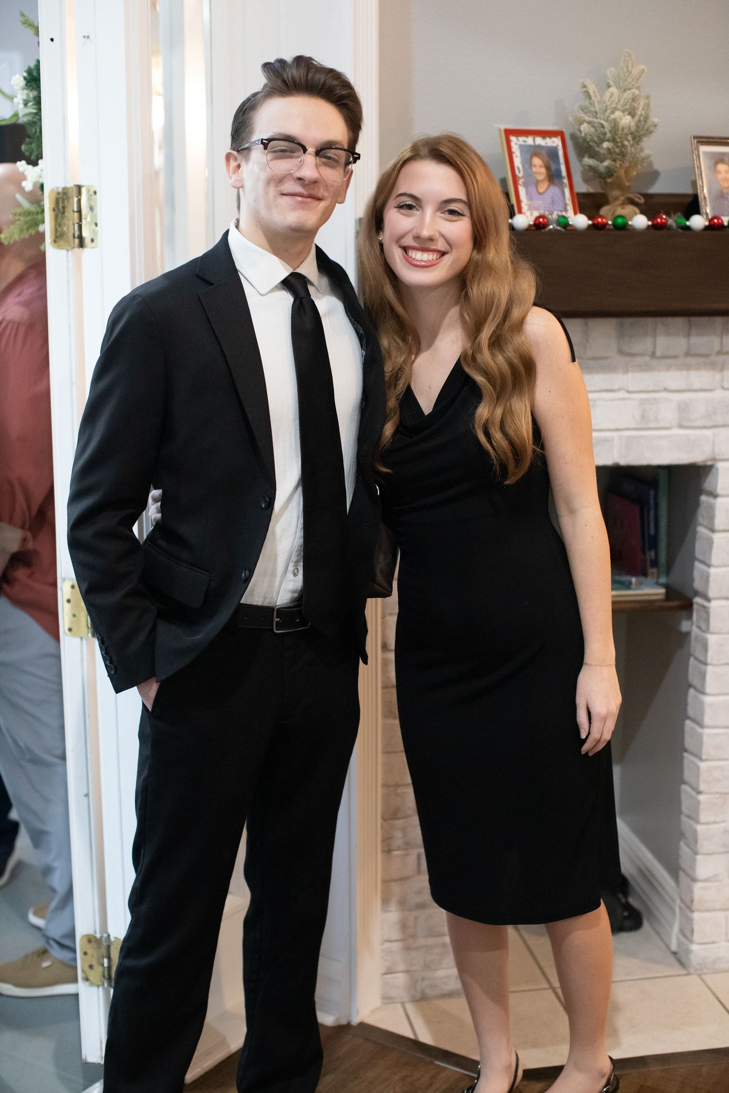 Couple in formal black attire posing by decorated fireplace at holiday party.