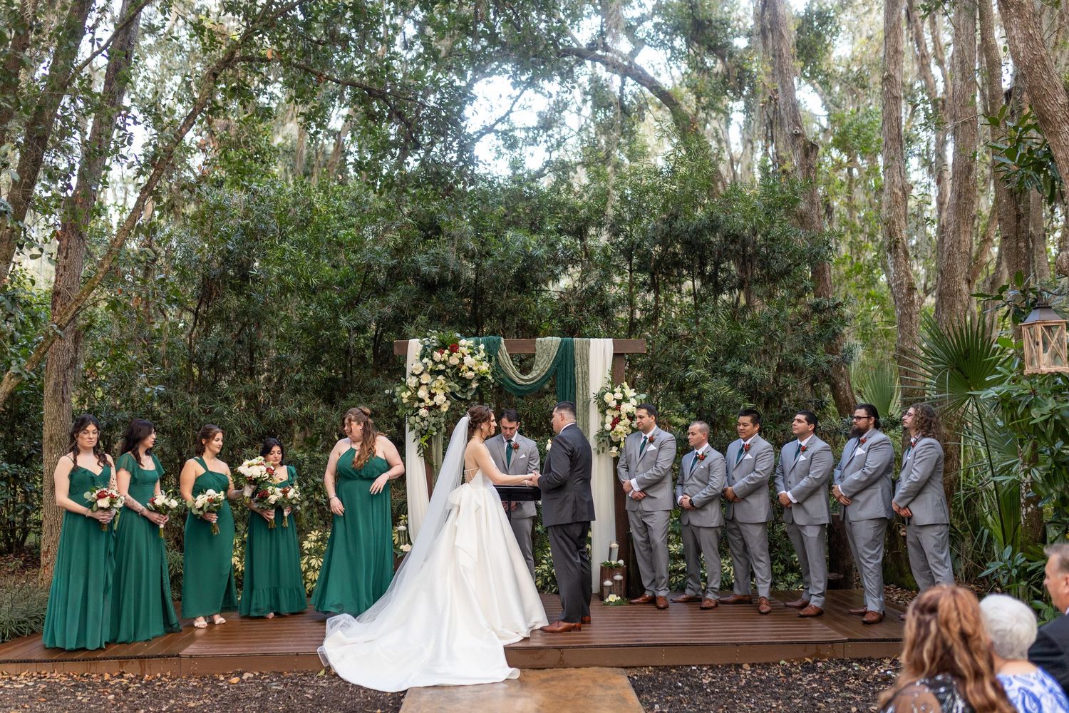 Wedding party in green and gray attire posed in a wooded outdoor setting.