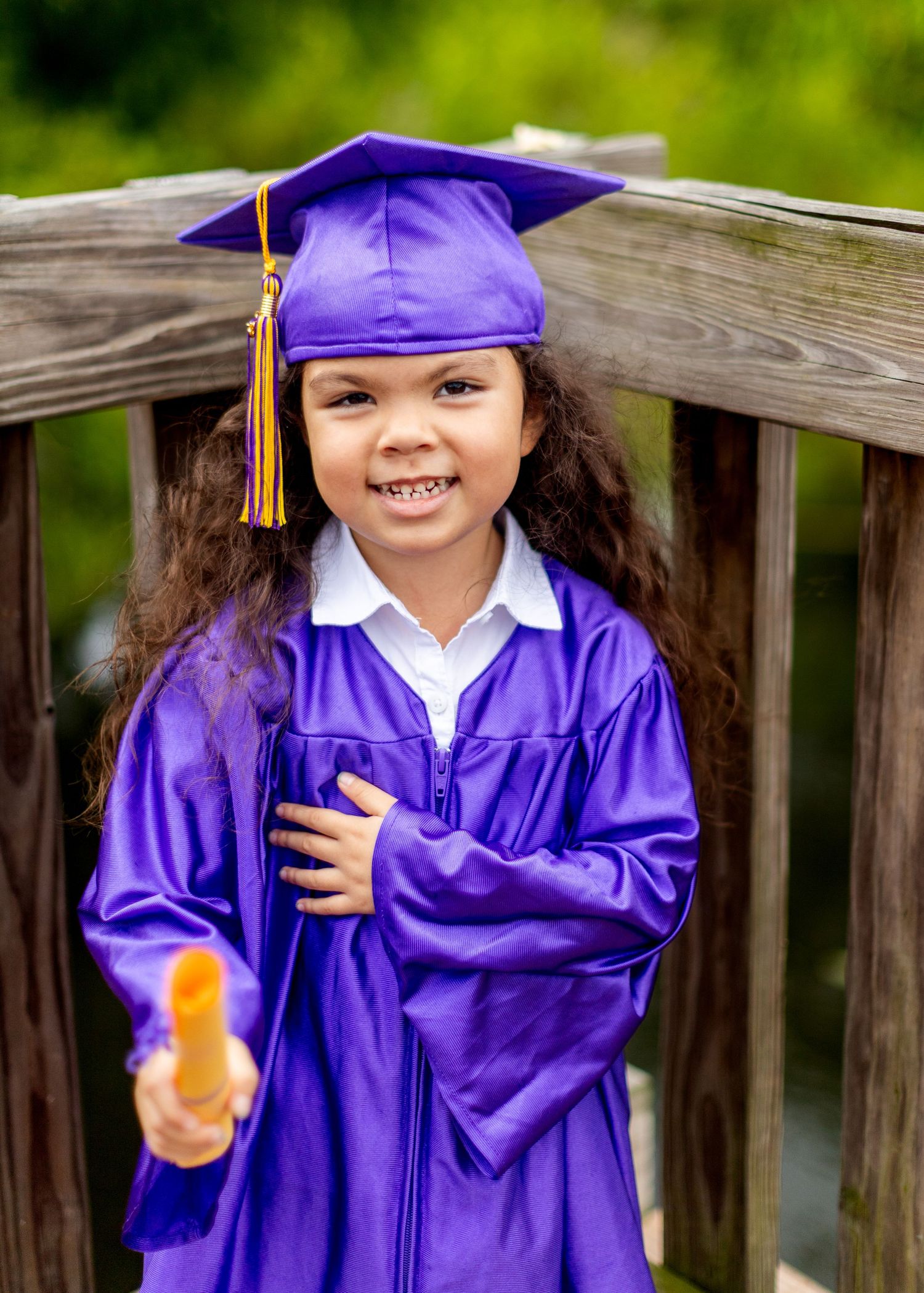Graduate in purple cap and gown holding diploma while standing on wooden deck.