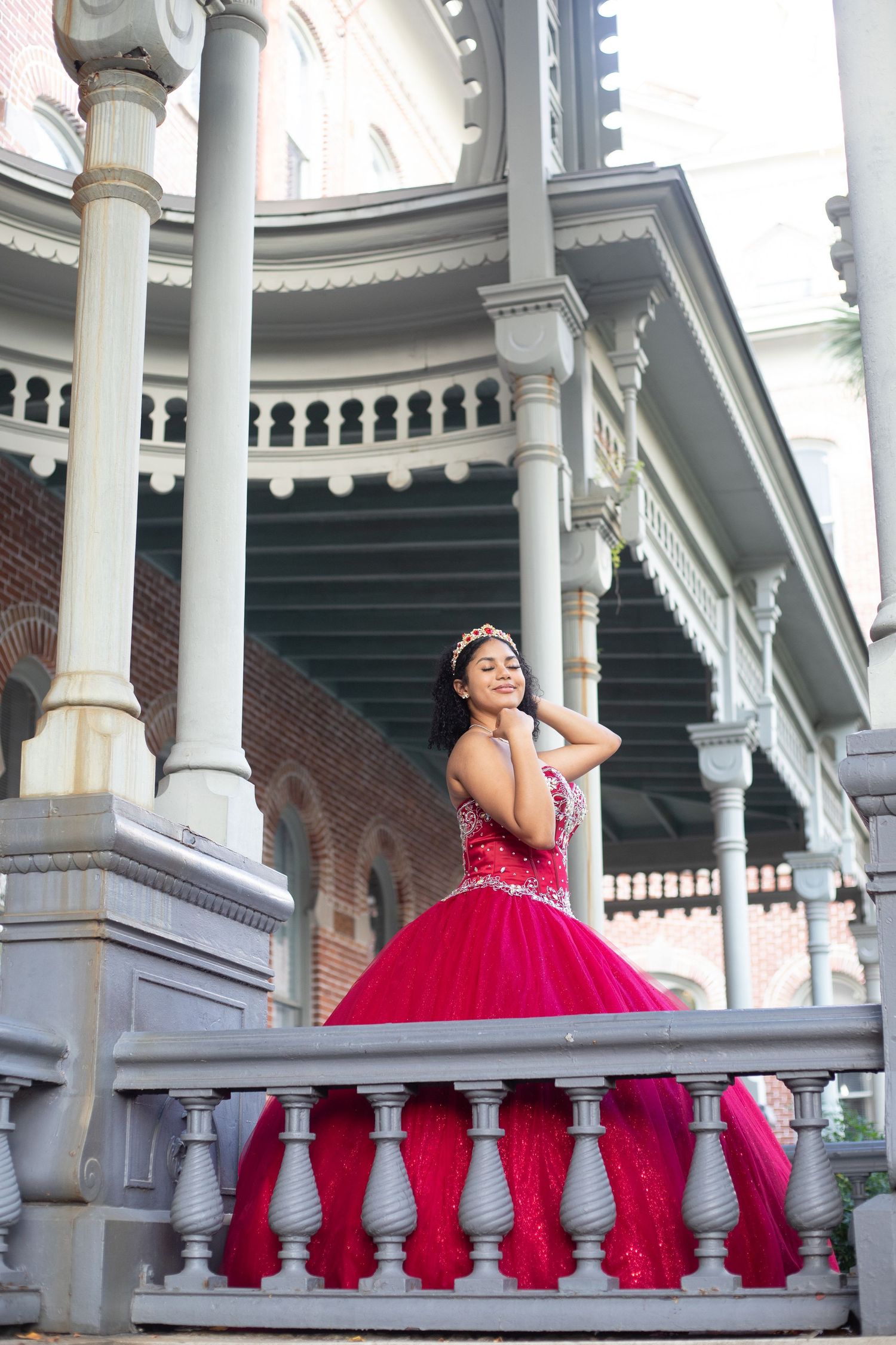 Elegant red ballgown photographed on white columned porch with ornate architecture.