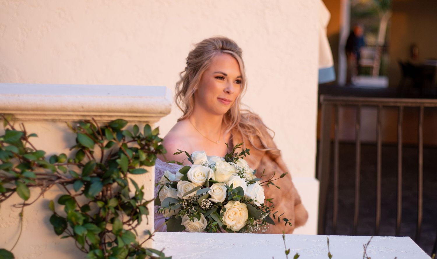 Elegant bride holding white rose bouquet in soft pink gown at outdoor wedding venue.