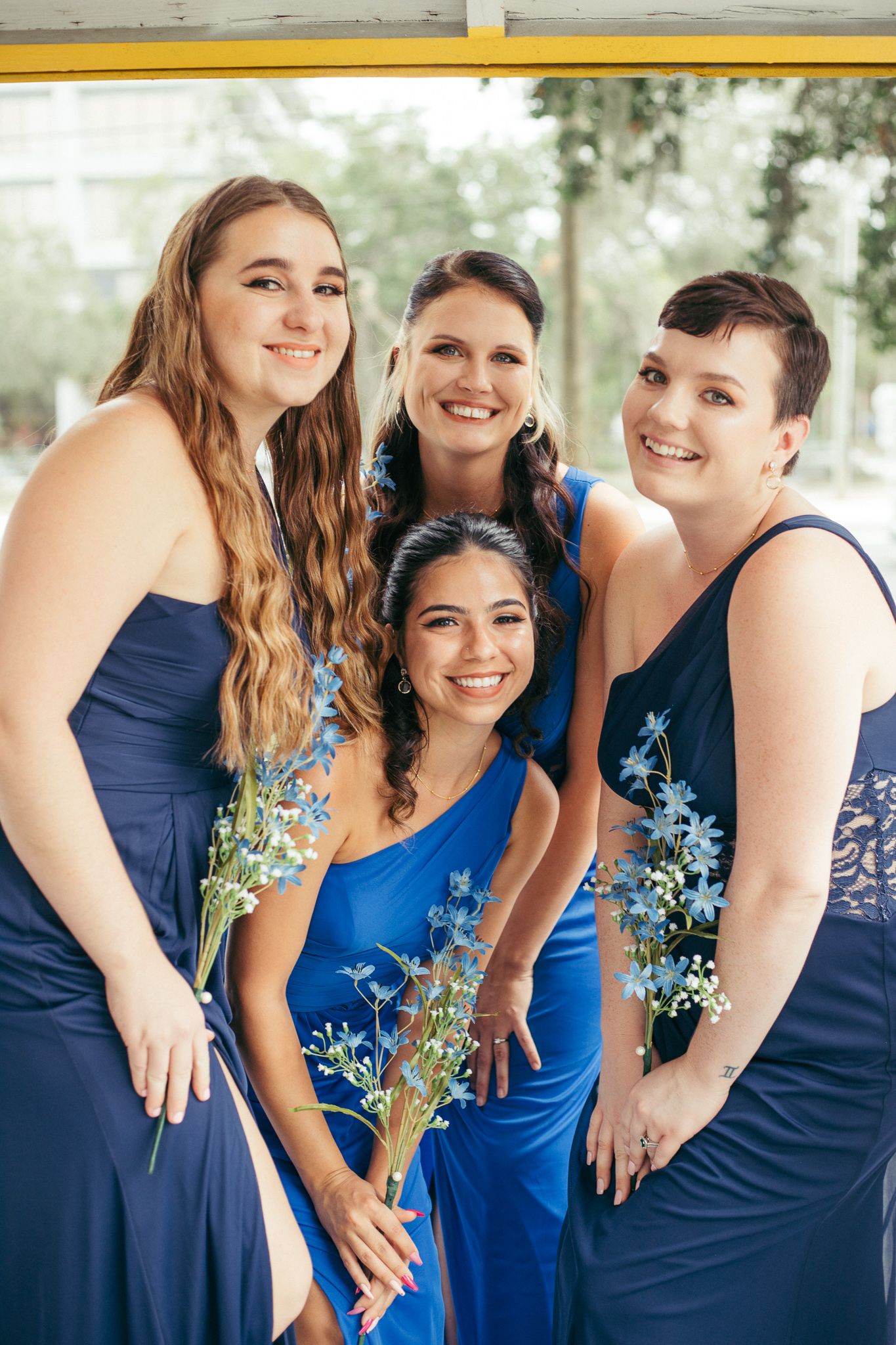 Four bridesmaids in navy blue dresses smiling and holding blue wildflowers at a wedding celebration.