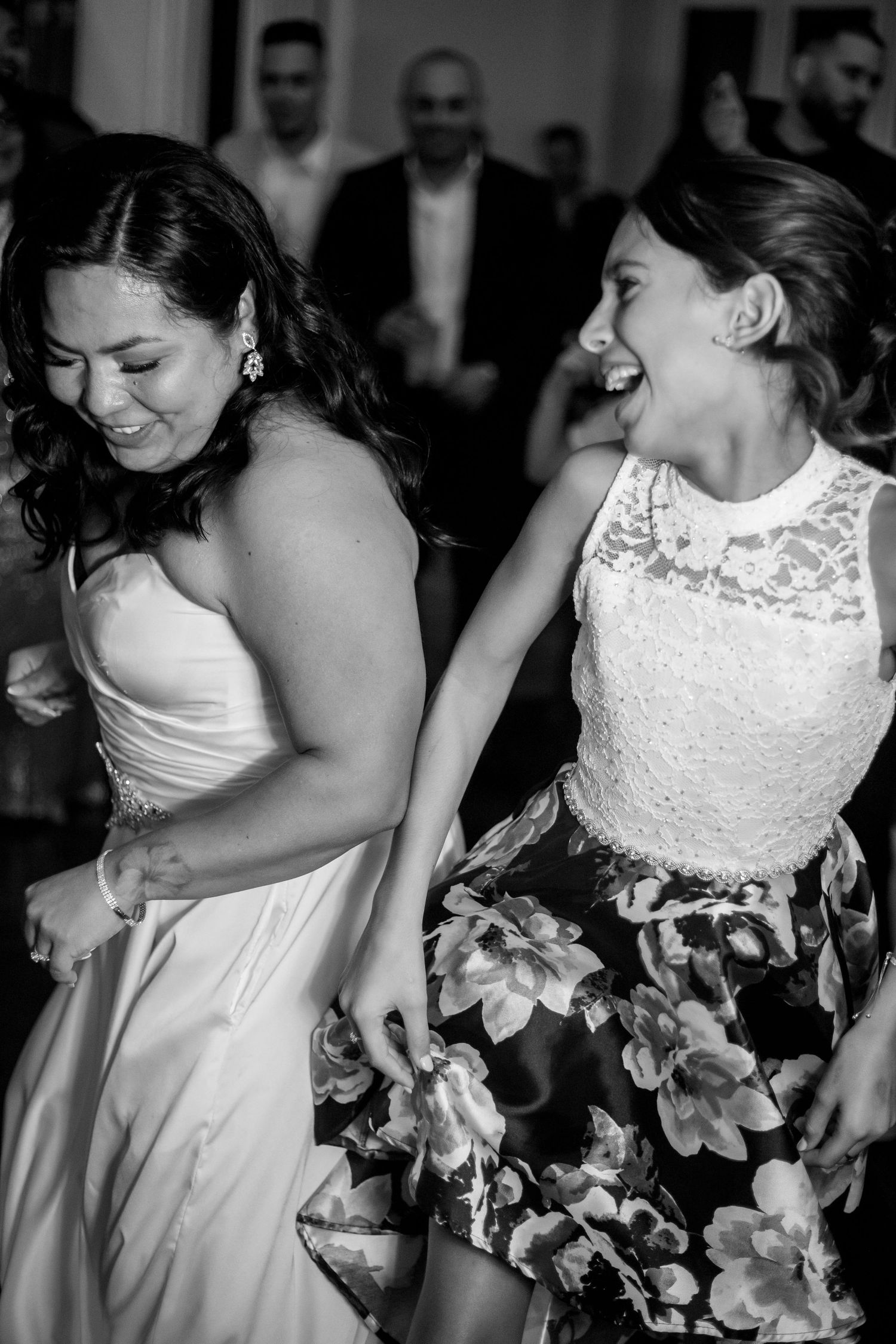 Two women laugh and dance joyfully at a wedding reception, one in a gown and one in a floral dress.