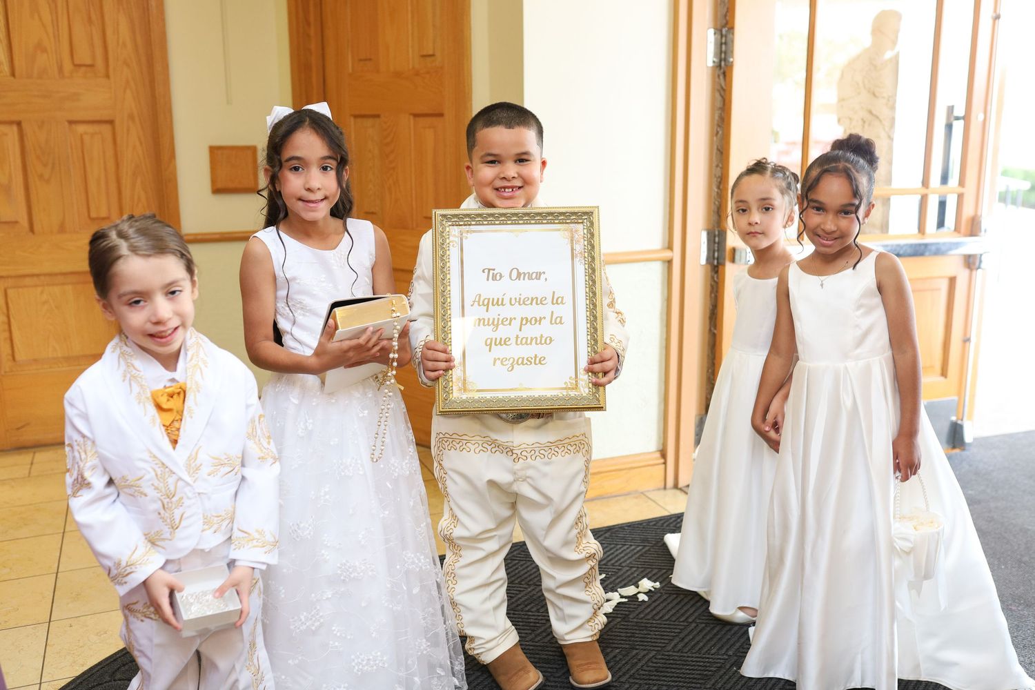 Five children in formal white attire pose together at a wedding ceremony, holding a framed sign in a church entrance.