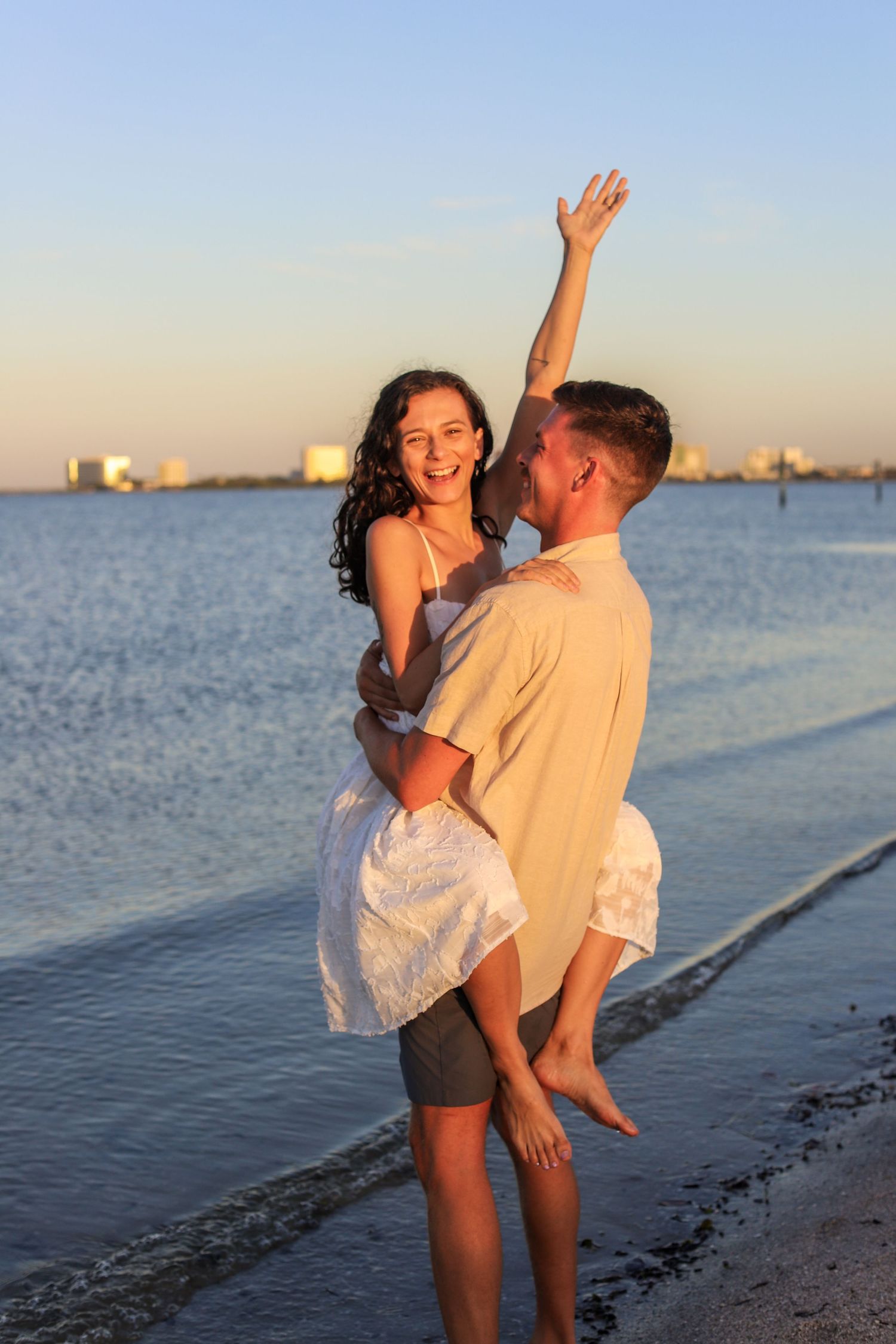 A joyful couple celebrates on a beach at sunset, man lifting woman in white dress as she raises her arm triumphantly.