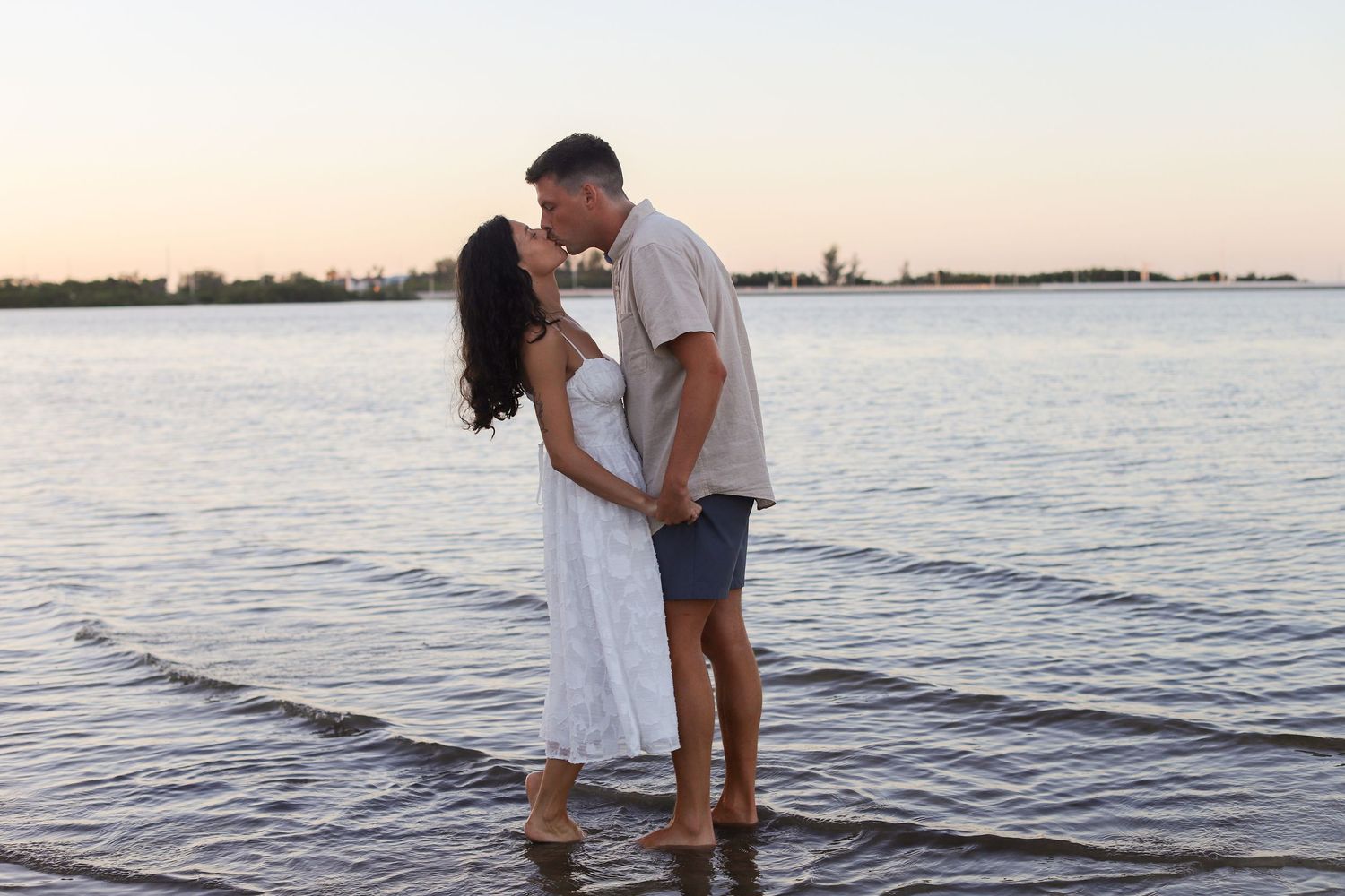 Couple kissing at sunset while standing in shallow water at the beach, woman in white dress, romantic engagement photo.