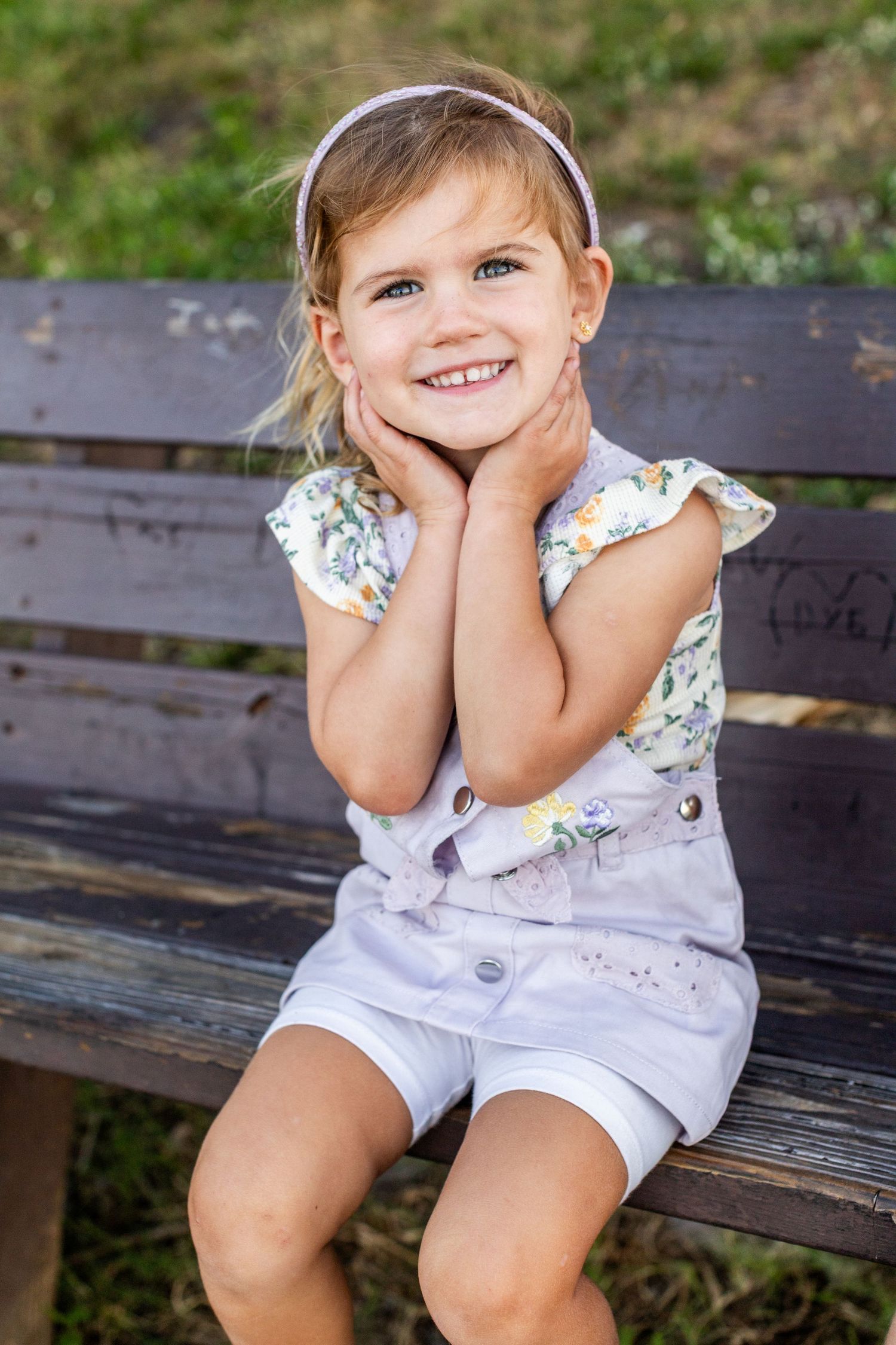 A cheerful little girl in a floral top and white shorts sits on a wooden bench, smiling with hands on cheeks.