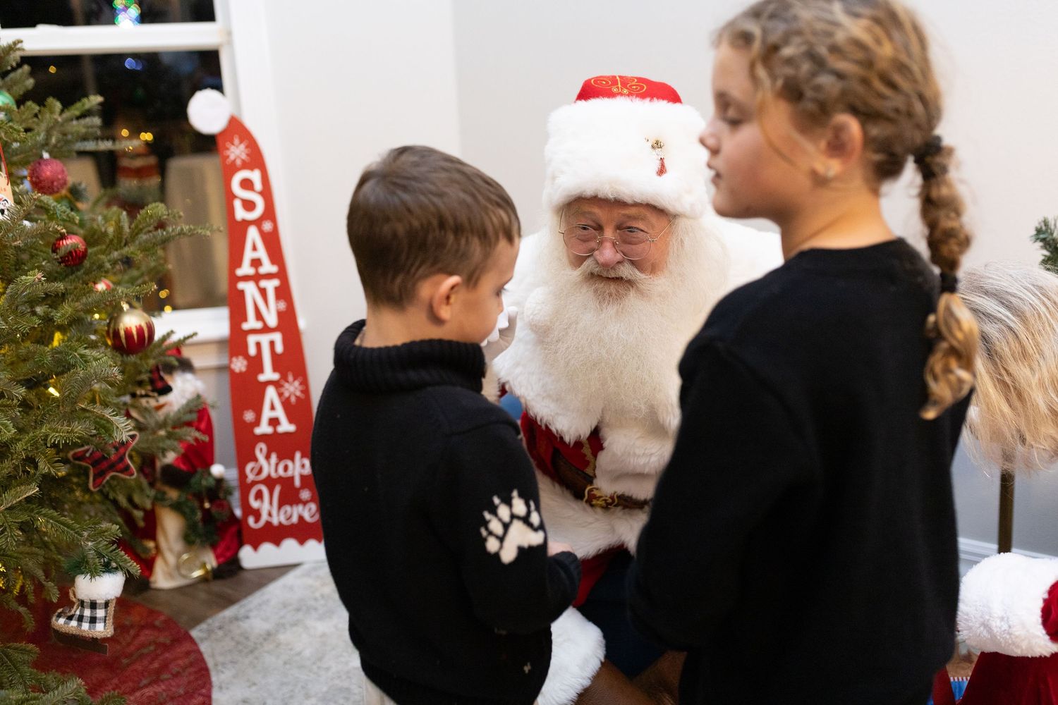 Santa Claus listens attentively during a Christmas meet-and-greet beside a decorated tree.