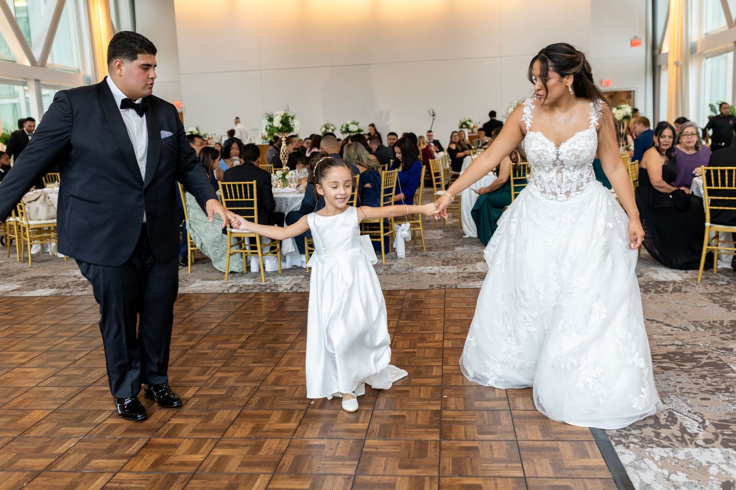Bride and groom dancing with flower girl in white dress at elegant wedding reception on wooden dance floor.