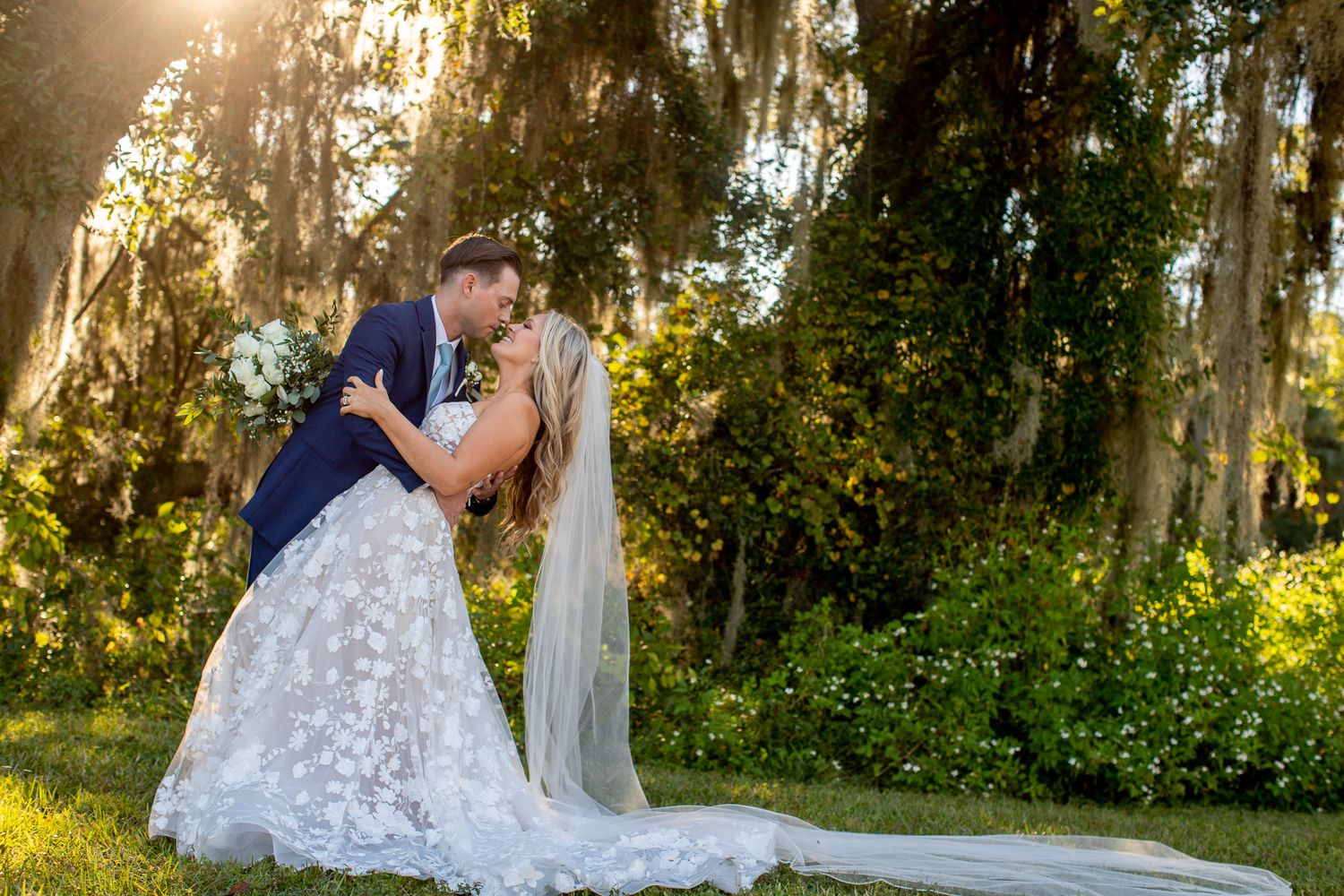 Bride and groom share a romantic dip kiss surrounded by moss-draped trees in golden sunset light.