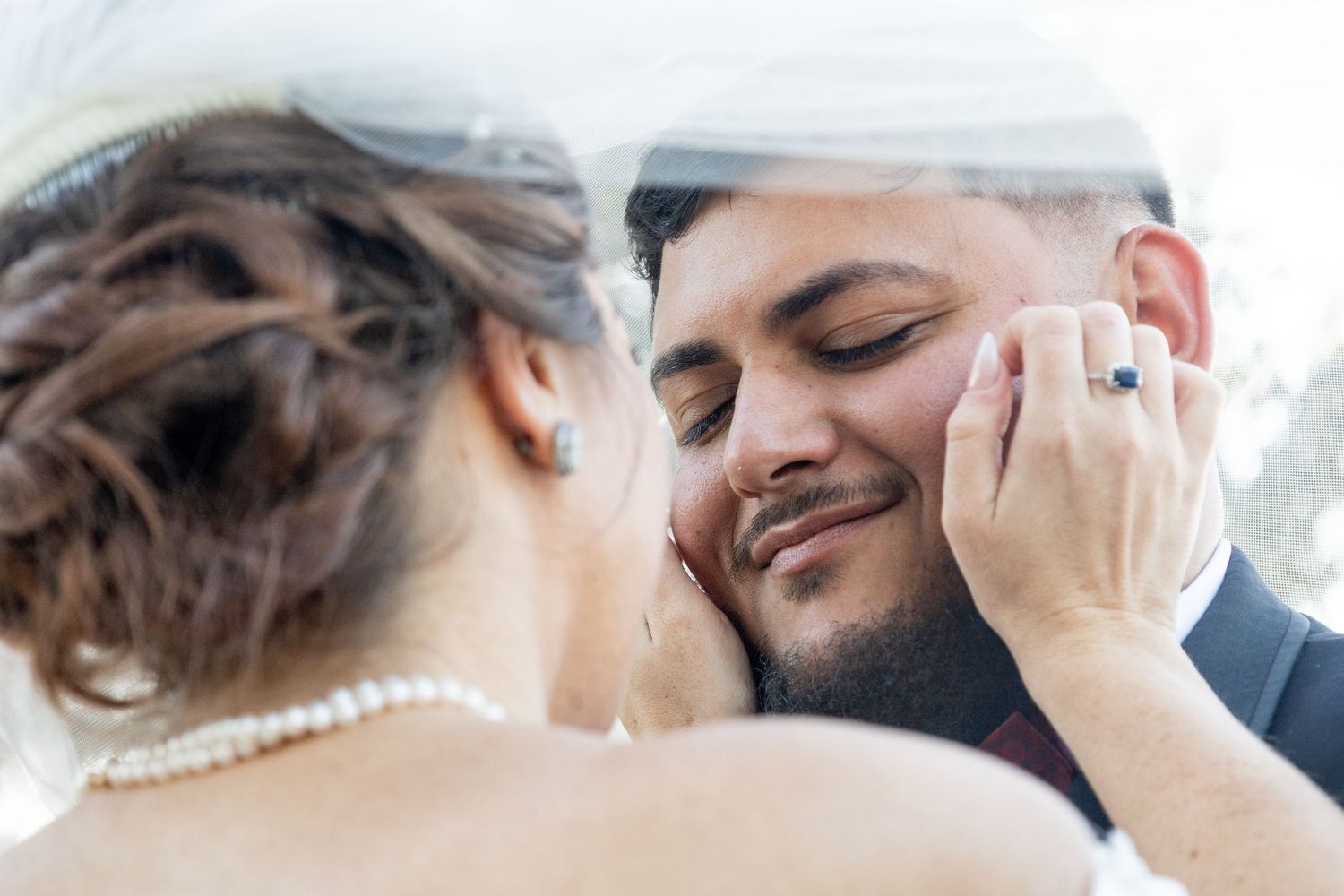 Bride and groom share an intimate moment under a veil, with bride wearing a sapphire engagement ring.
