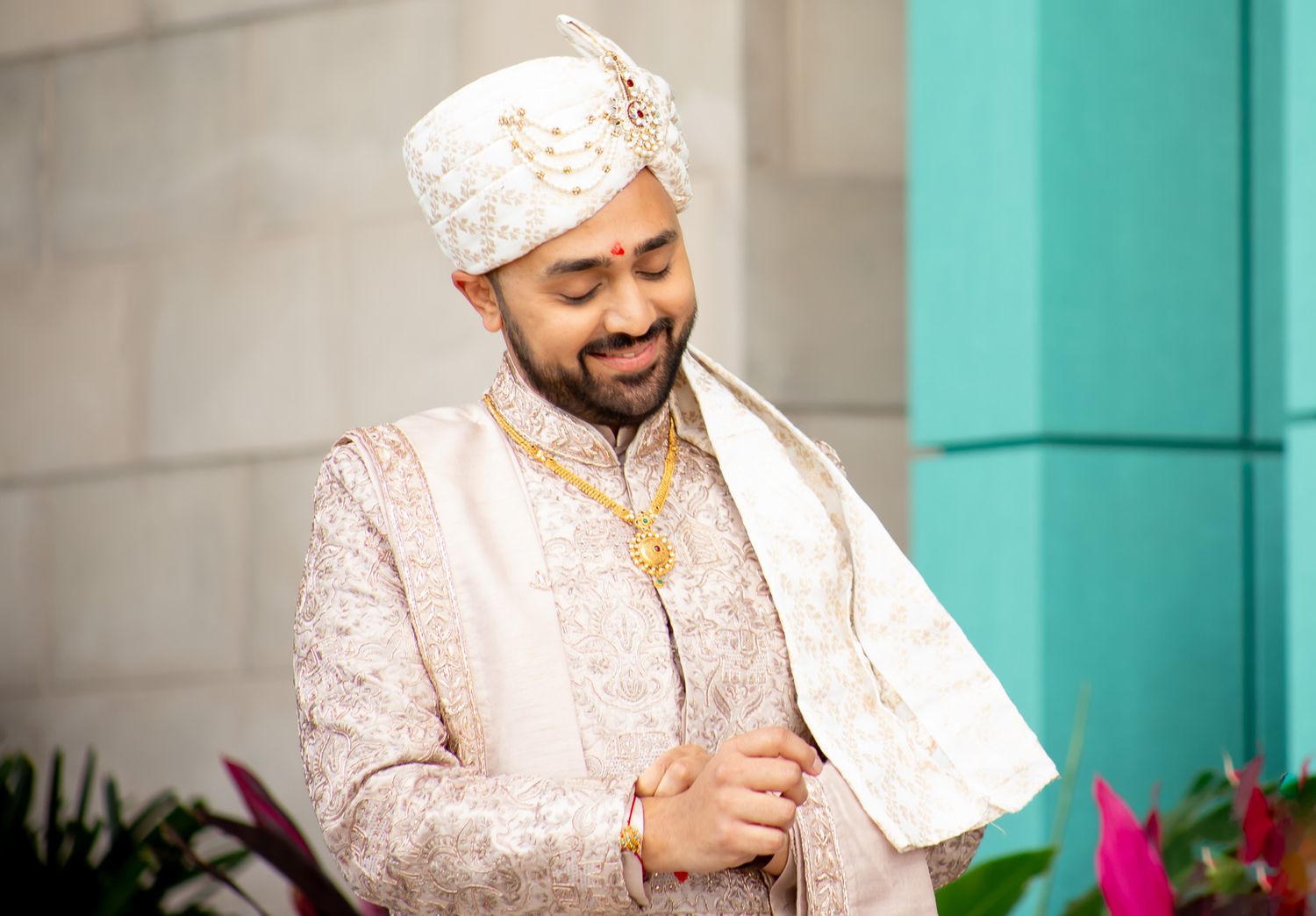 A groom in cream sherwani and turban smiles against a turquoise wall with pink flowers.