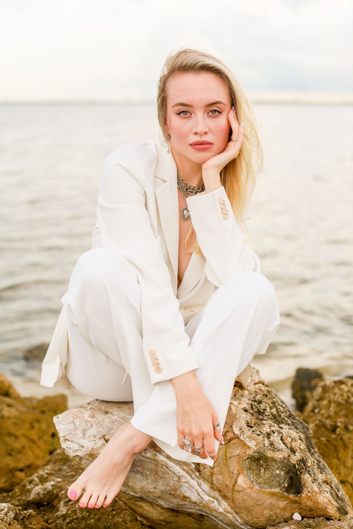 Elegant coastal portrait featuring white suit and statement jewelry against ocean backdrop.