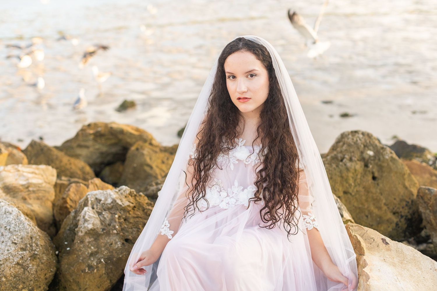 A dreamy beach portrait featuring flowing white lace and curls against rocky coastal backdrop at sunset.