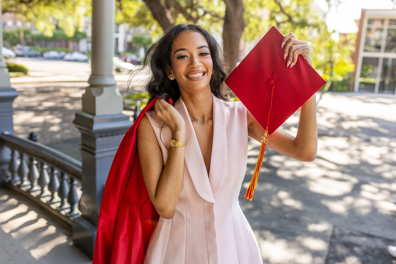 Smiling young woman in pink dress holding red graduation cap and gown outdoors on a sunny day.
