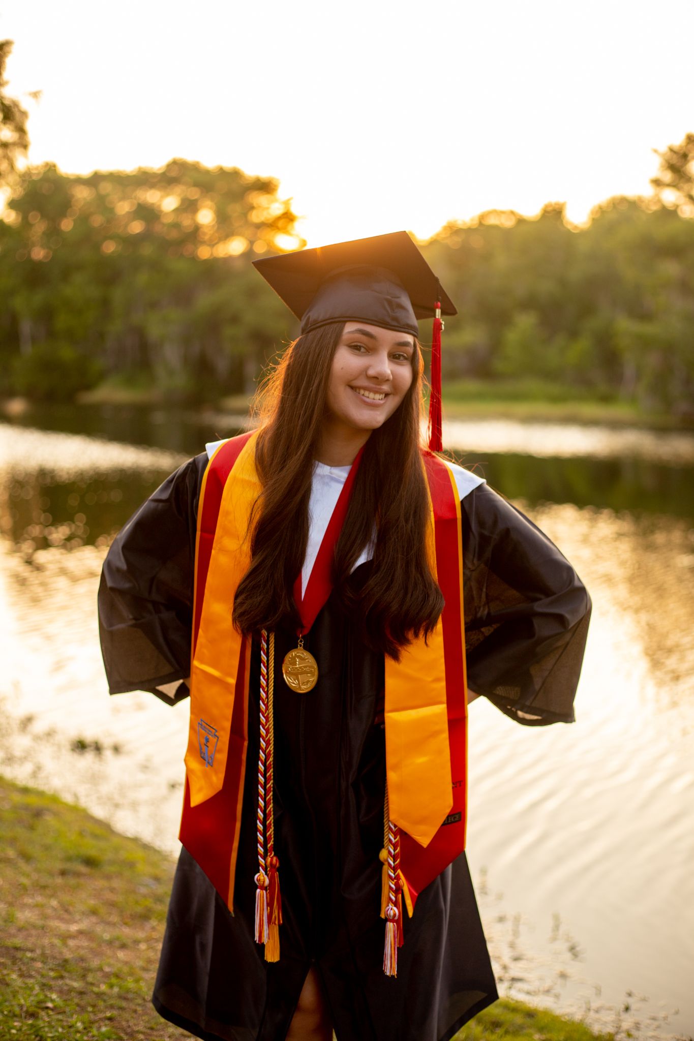 Graduate in black gown and gold sash poses at sunset by a lake.