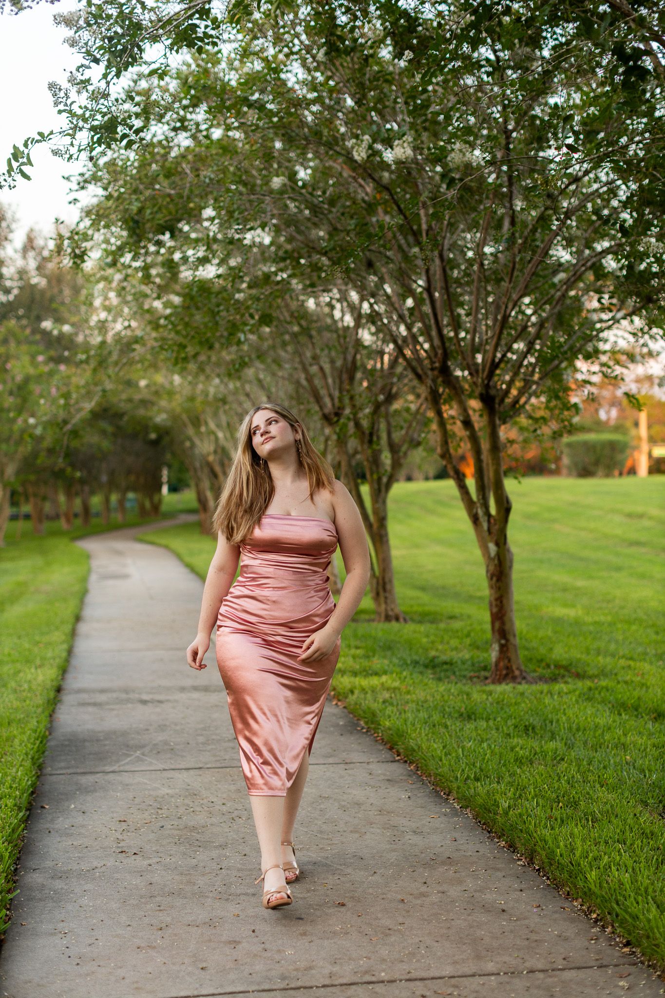 Flowing pink dress walking along tree-lined pathway in park setting.