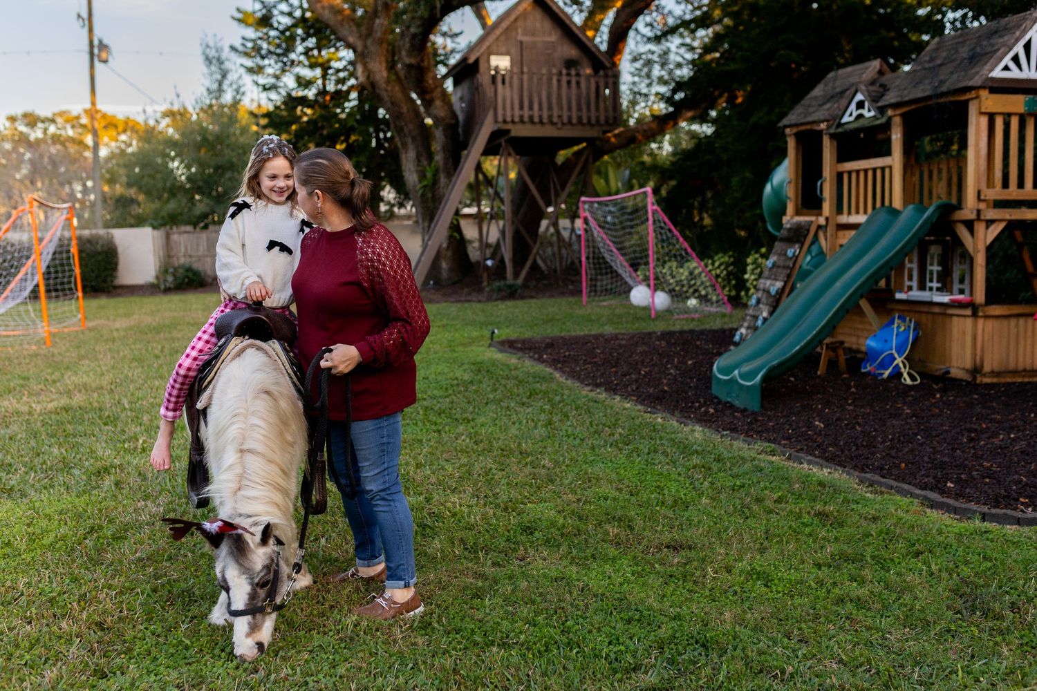 A white pony with a saddle stands in a grassy backyard area near a wooden playground structure at sunset.