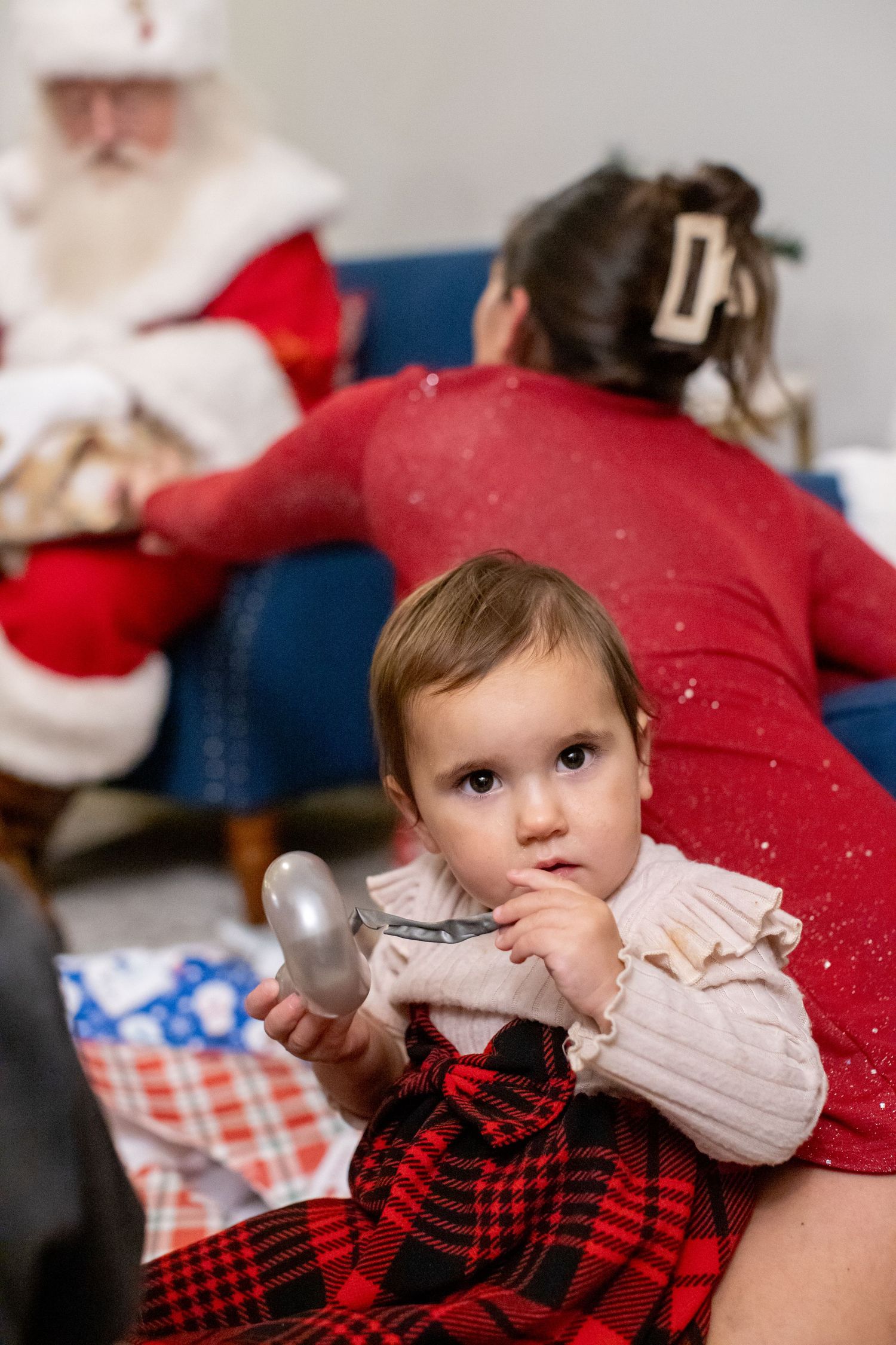 A baby in a red and black plaid dress rests on a couch during a holiday gathering.