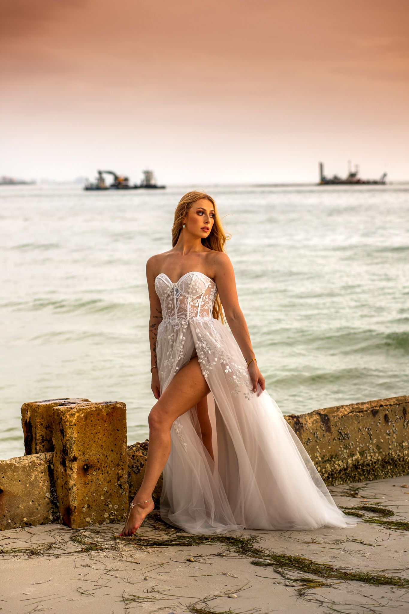 Bride in flowing white gown poses gracefully on beach at sunset with ships in background.