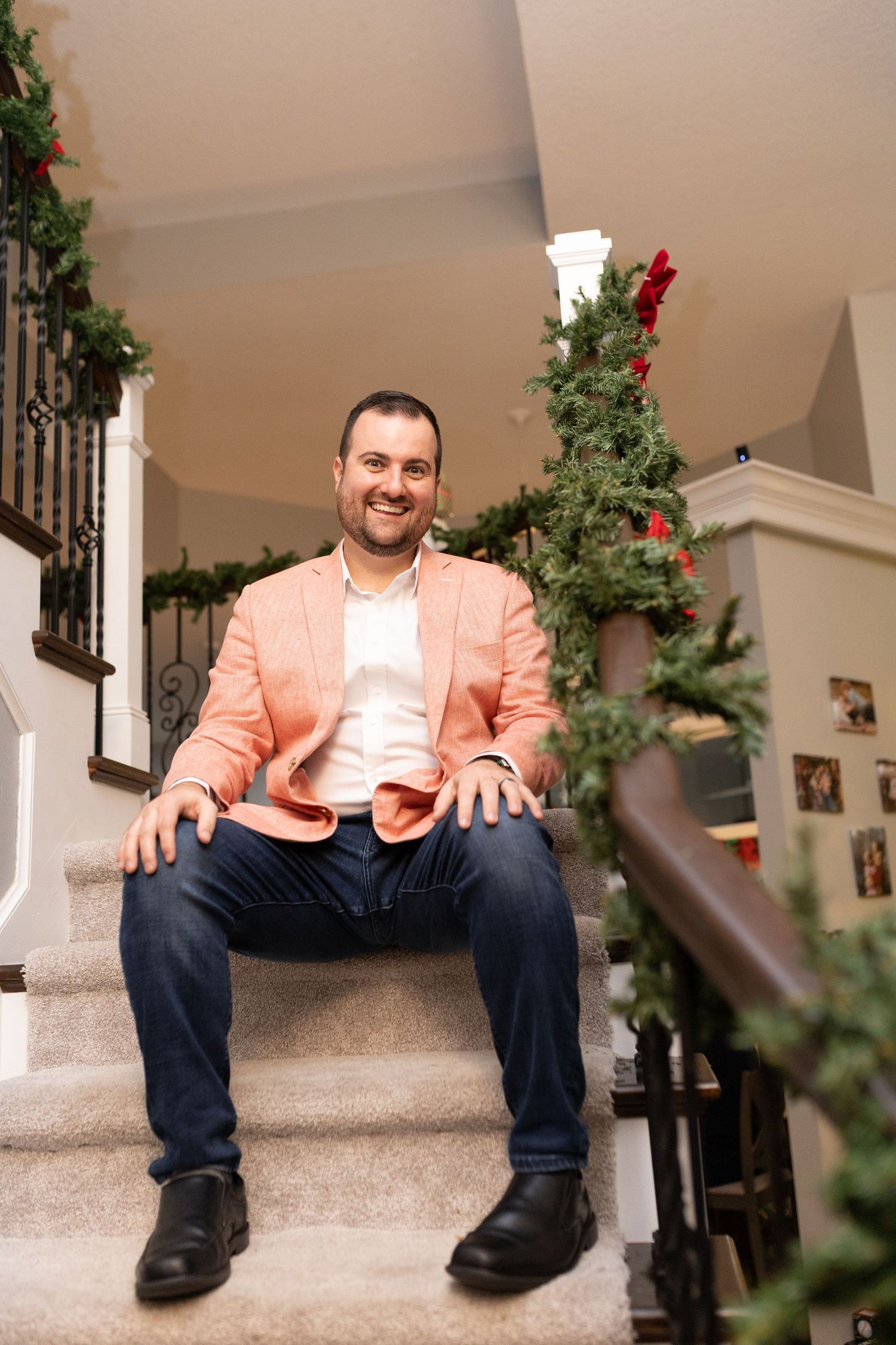 Someone wearing a peach-colored blazer and jeans sits on carpeted stairs decorated with holiday greenery.