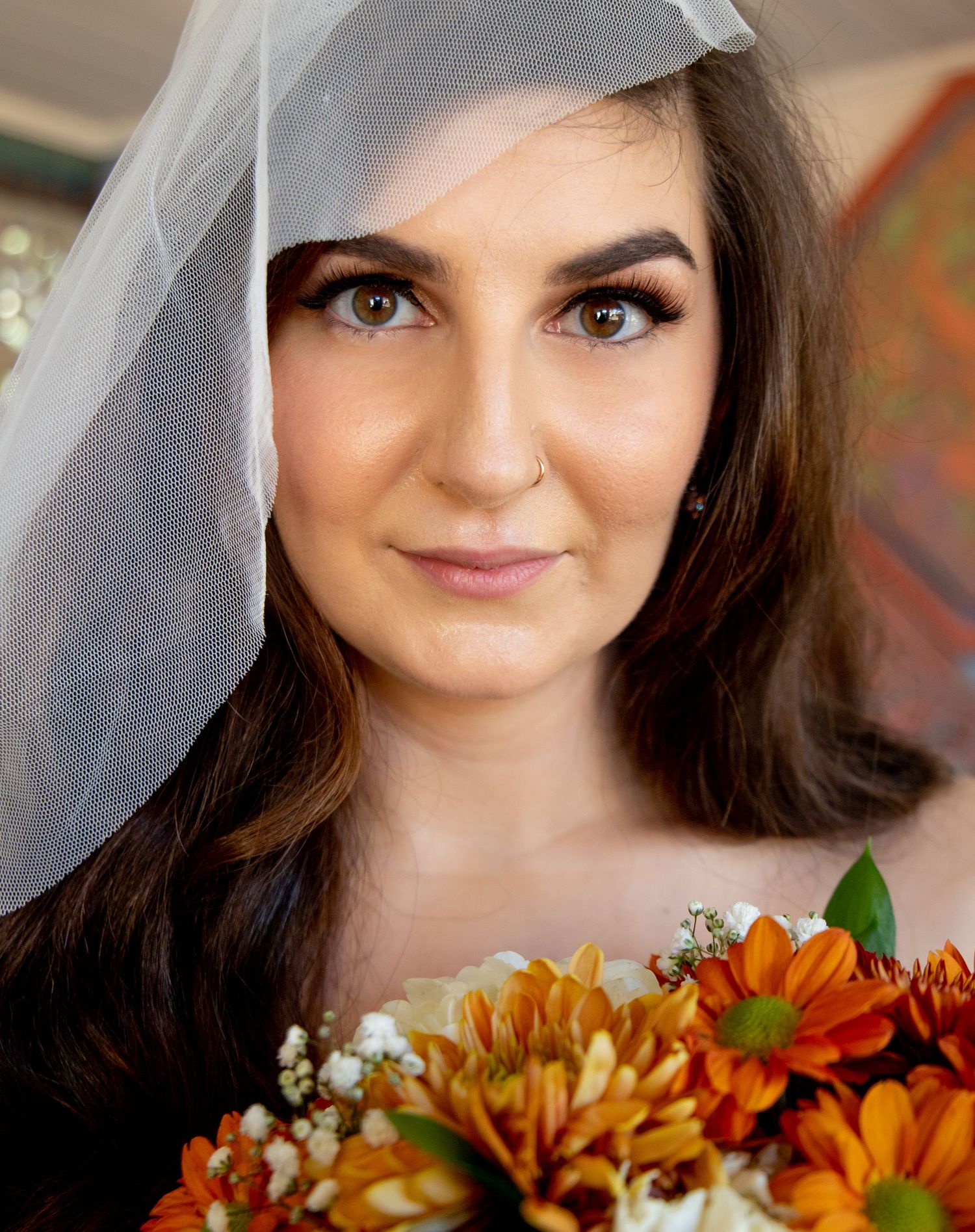 A beautiful bride wearing a white veil holds an orange floral bouquet, with natural makeup and dark hair.
