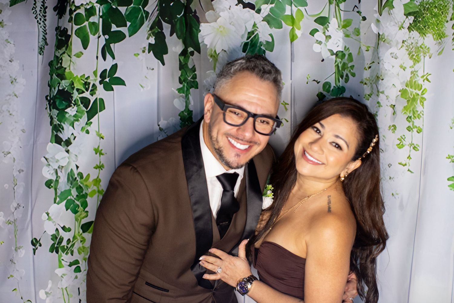 A smiling couple in formal wedding attire poses together under a floral arch with white drapes and green leaves.