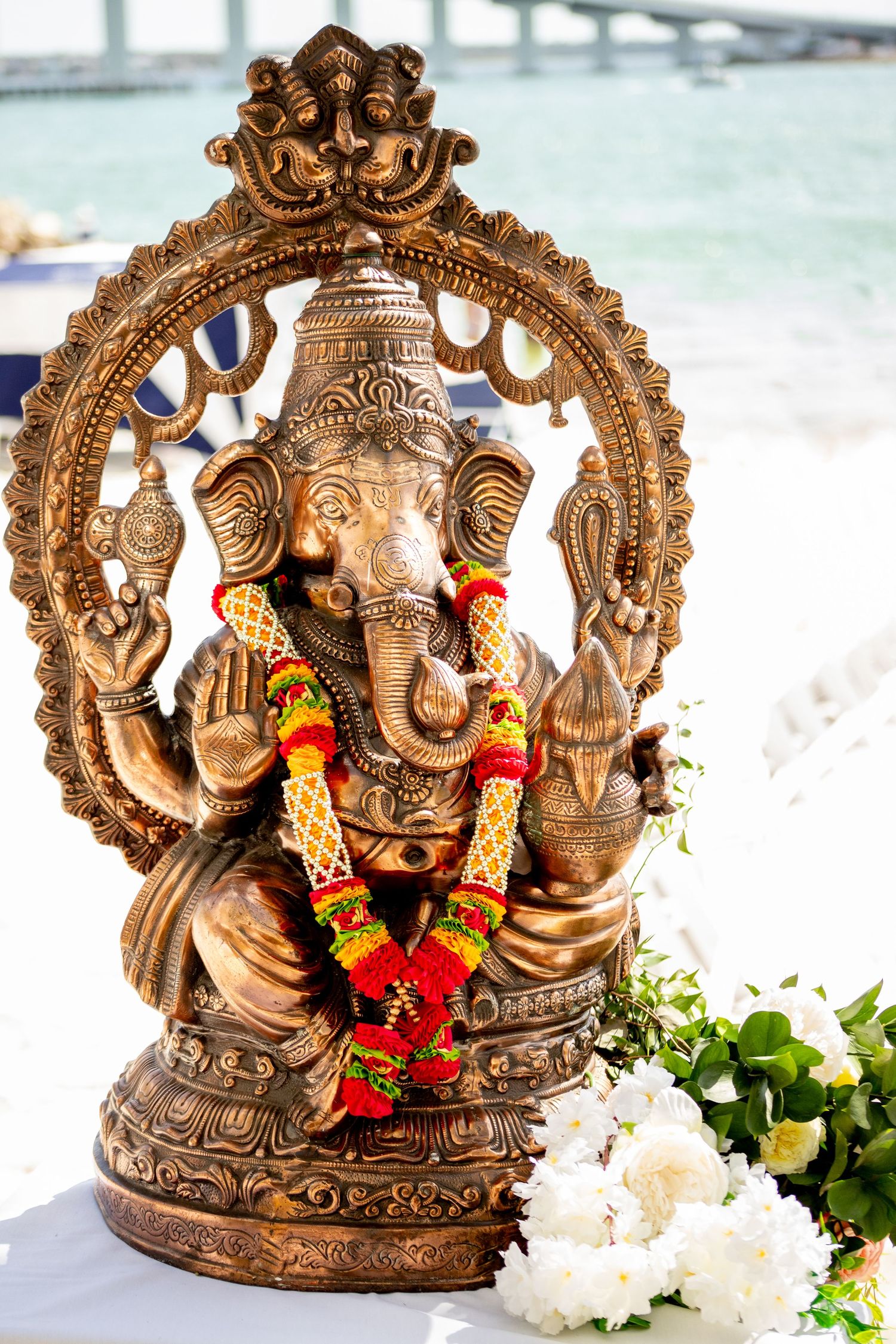 Bronze Ganesha statue adorned with colorful floral garland, surrounded by white flowers, near a waterfront setting.