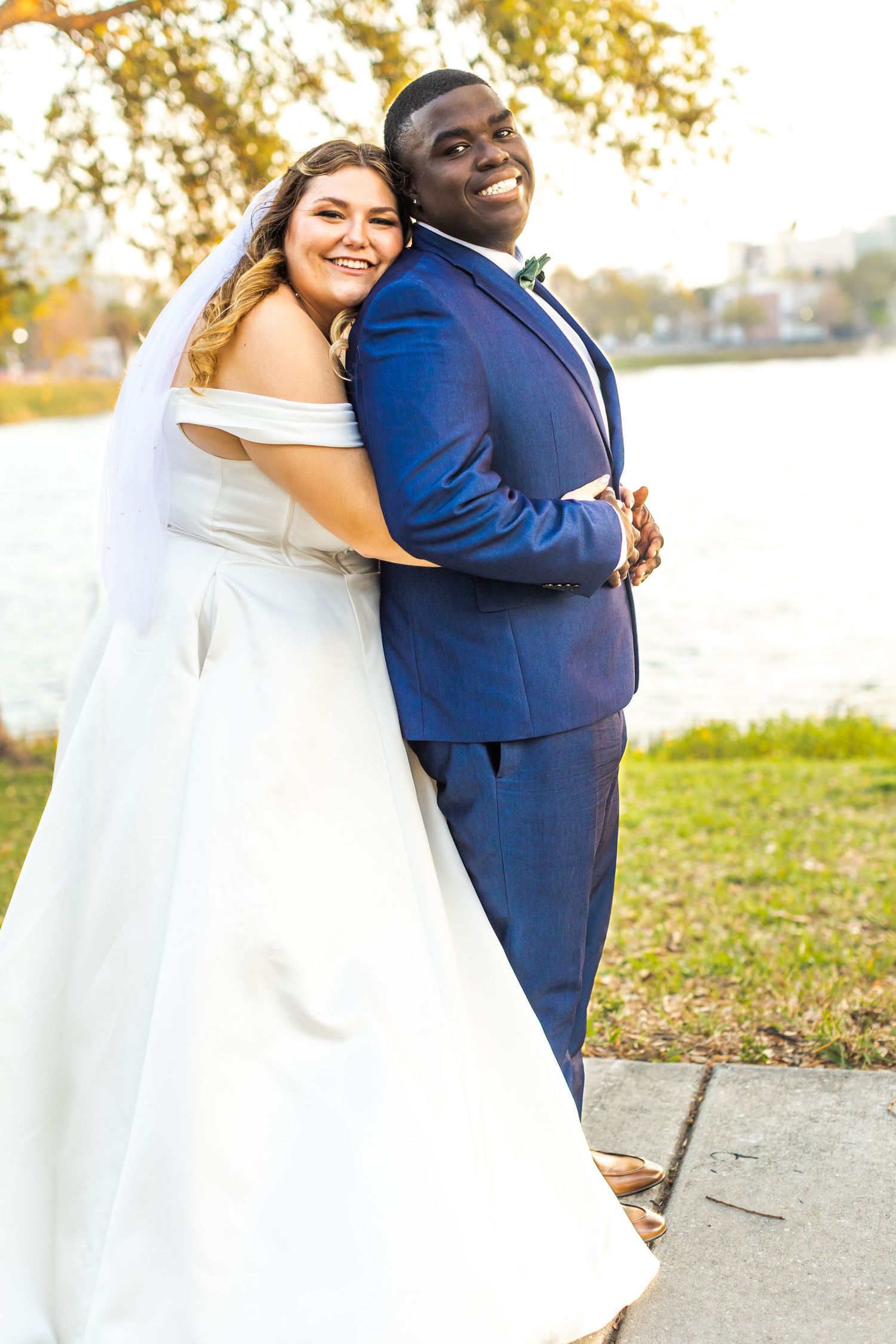 A happy couple in wedding attire poses back-to-back by a waterfront.