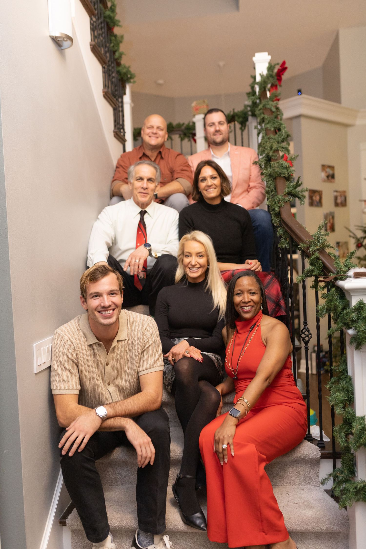 People in formal attire gathered on carpeted stairs decorated with holiday greenery for a group photo.