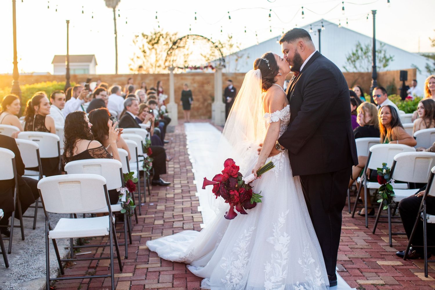Bride and groom share a kiss at outdoor wedding ceremony with white chairs and red bouquet at sunset.