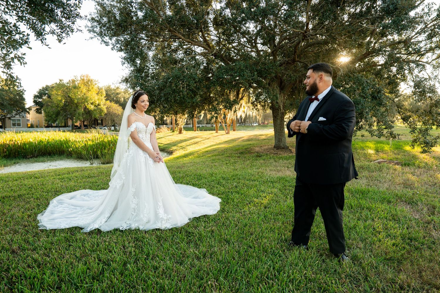 Bride in white ball gown and groom in black tuxedo share first look moment on lush green lawn under oak trees at sunset.
