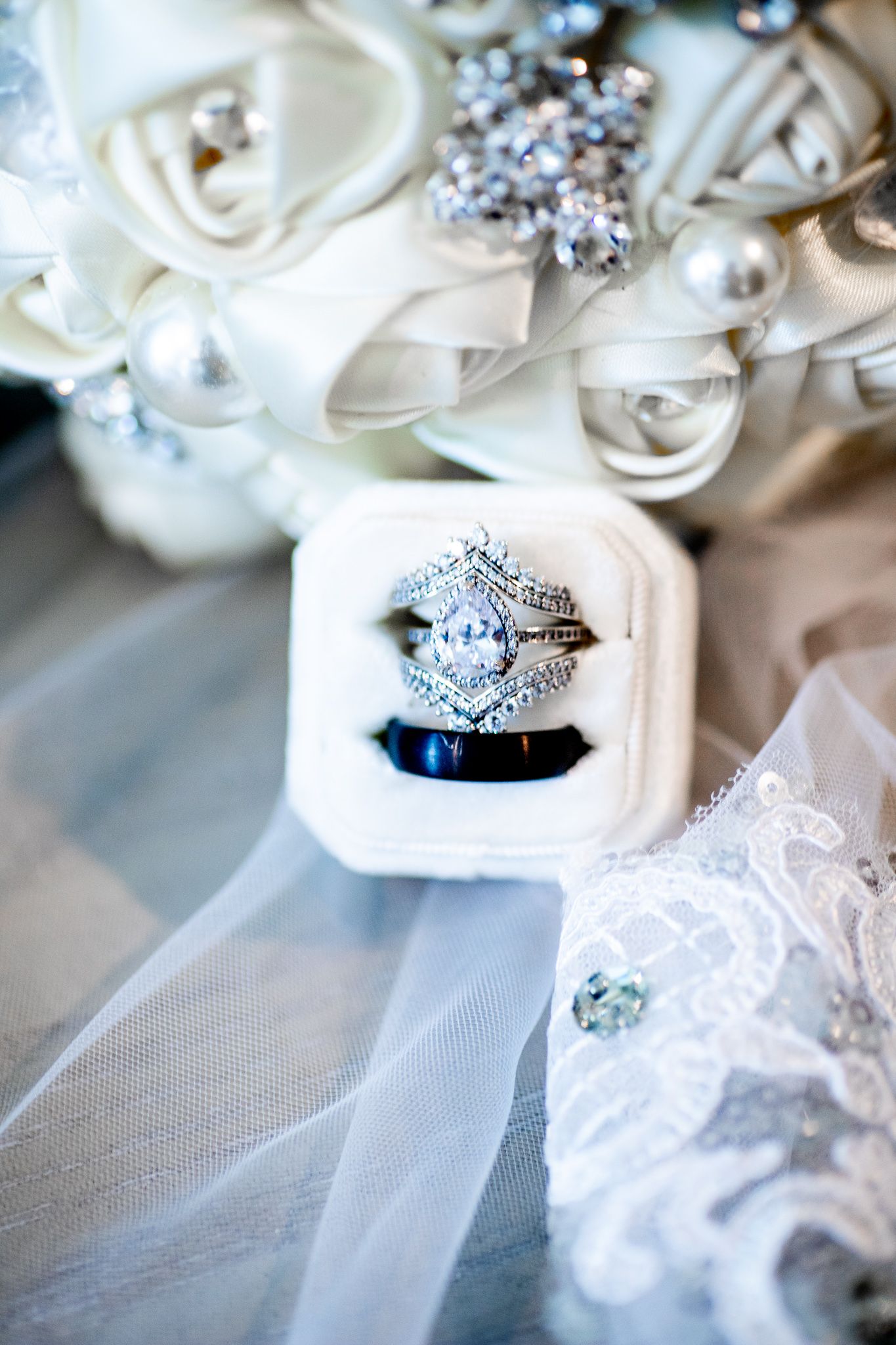 Elegant diamond engagement ring and wedding bands displayed in a white velvet box with bridal bouquet and lace veil.