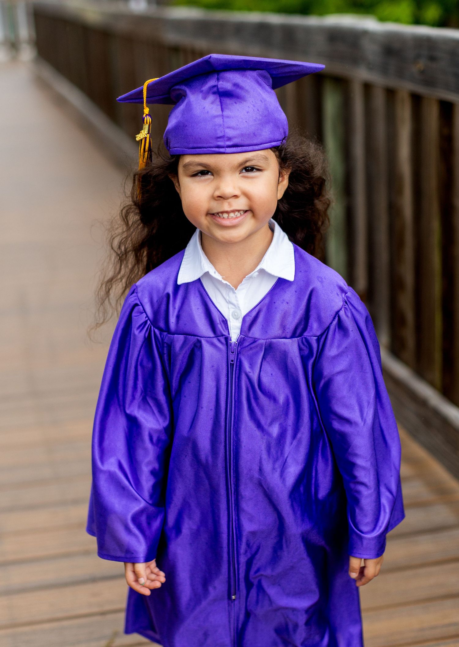 Graduate in vibrant purple cap and gown smiling while walking on wooden deck.
