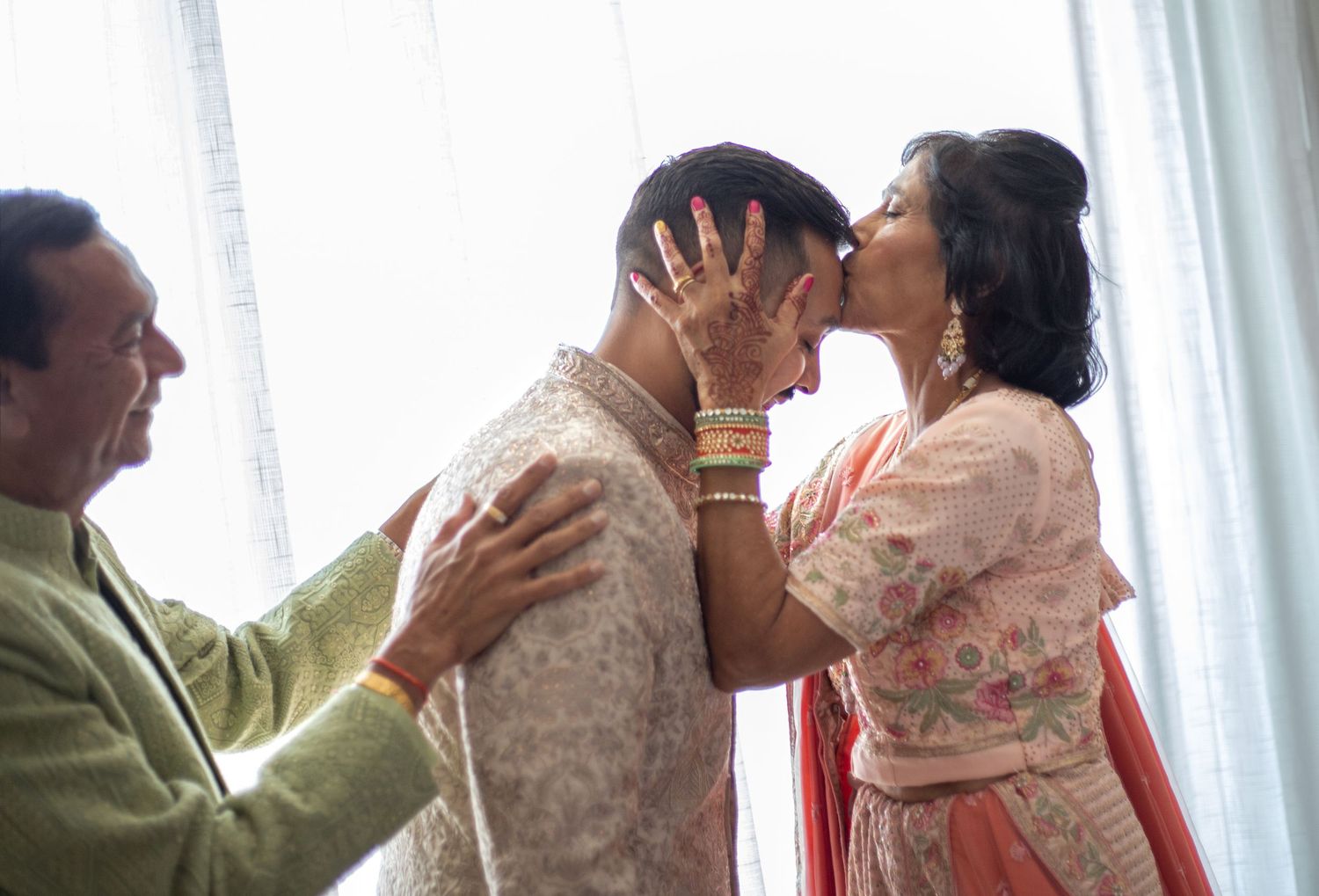 A woman kisses a young man in traditional Indian attire while an older man places his hand on his shoulder.