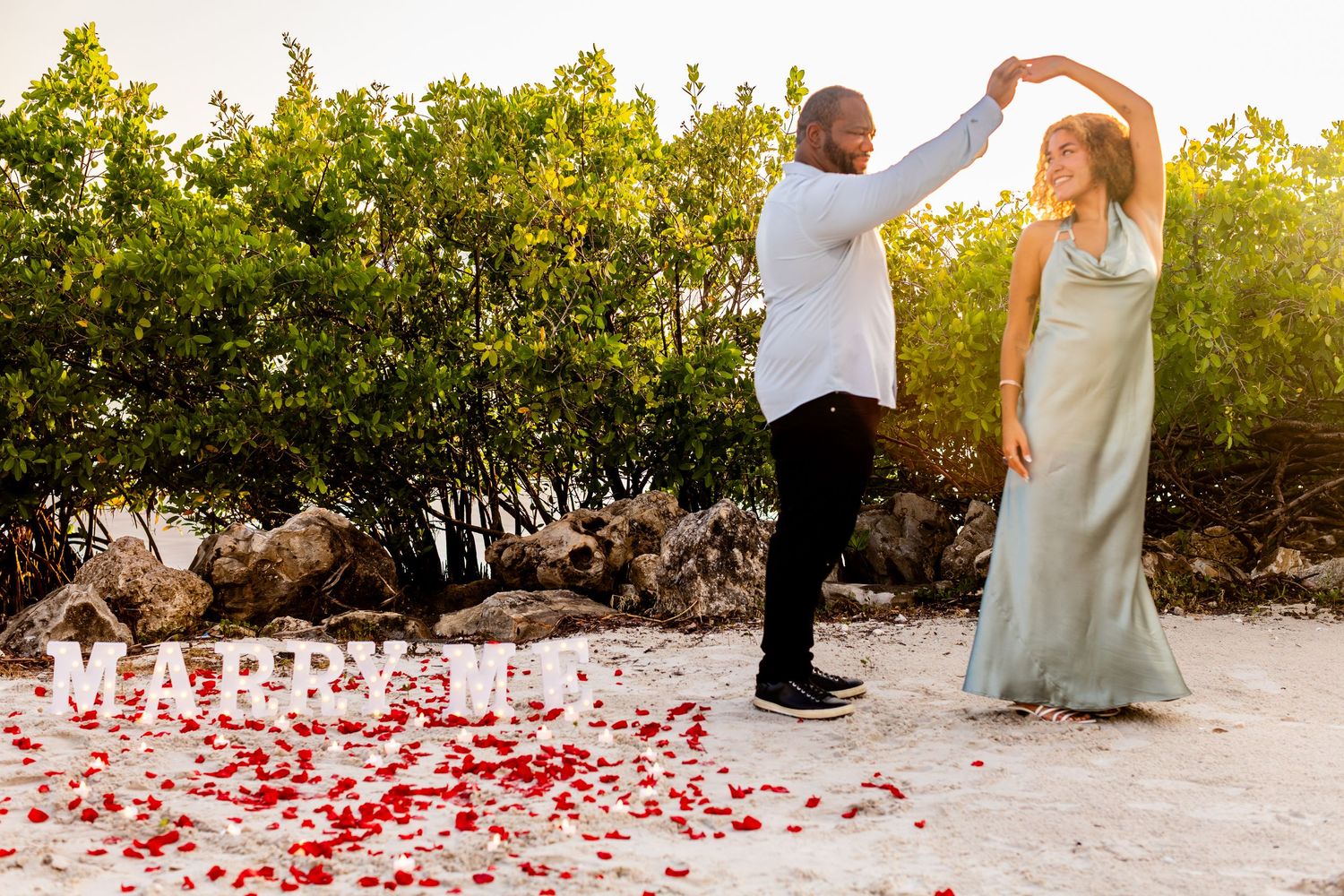 Couple dancing on beach with rose petals and MARRY ME sign during romantic outdoor marriage proposal.